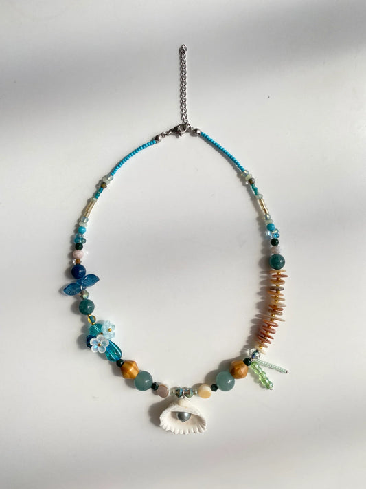 Flat lay shot on a white table of the handcrafted 1-of-1 blue necklace, clearly displaying the 3D bead flowers, shell spacers, and pendant details.