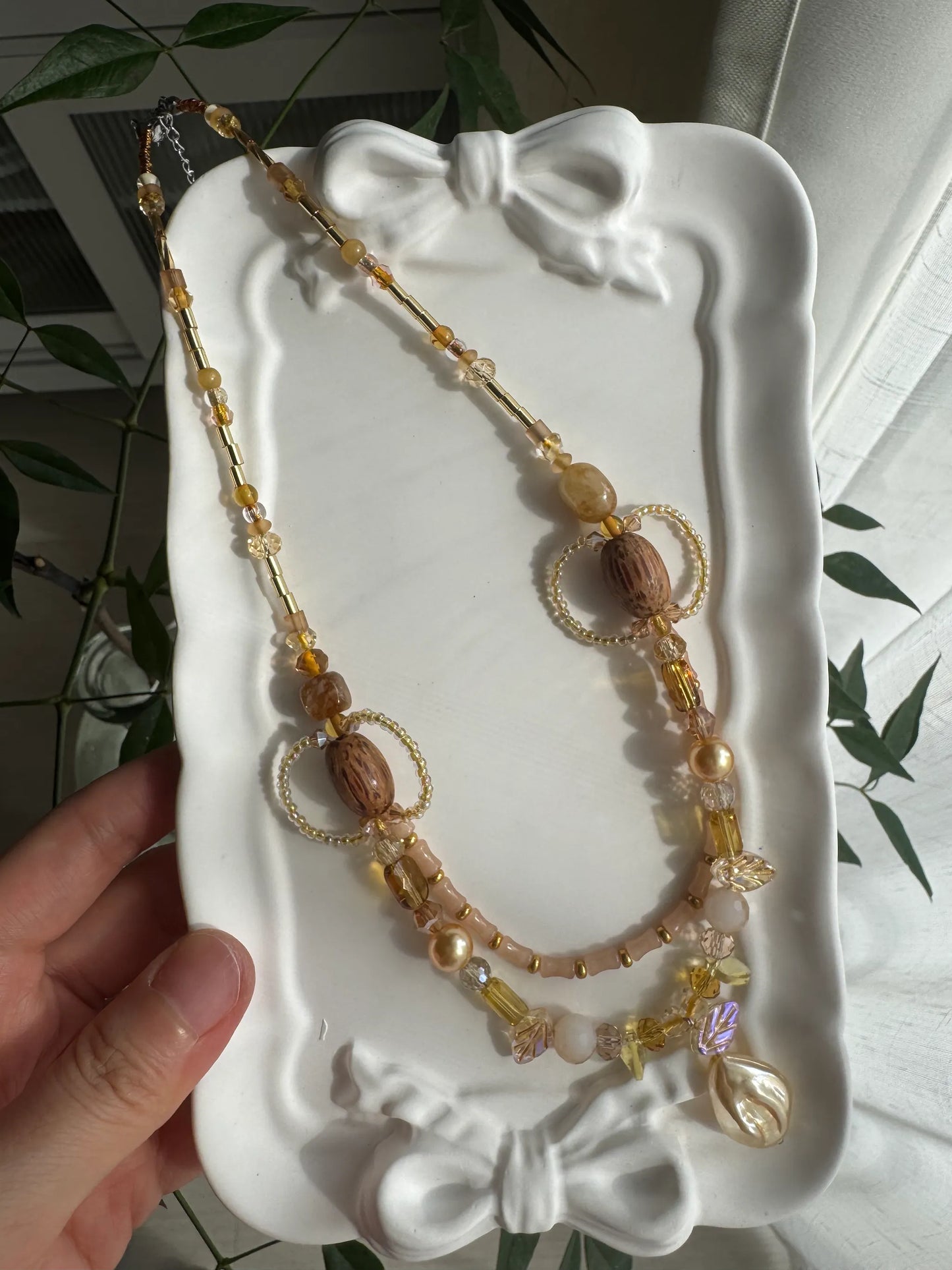 HuibenCraft 1-of-1 shimmering layered necklace with light yellow and brown beads in a white ceramic dish under natural sunlight.
