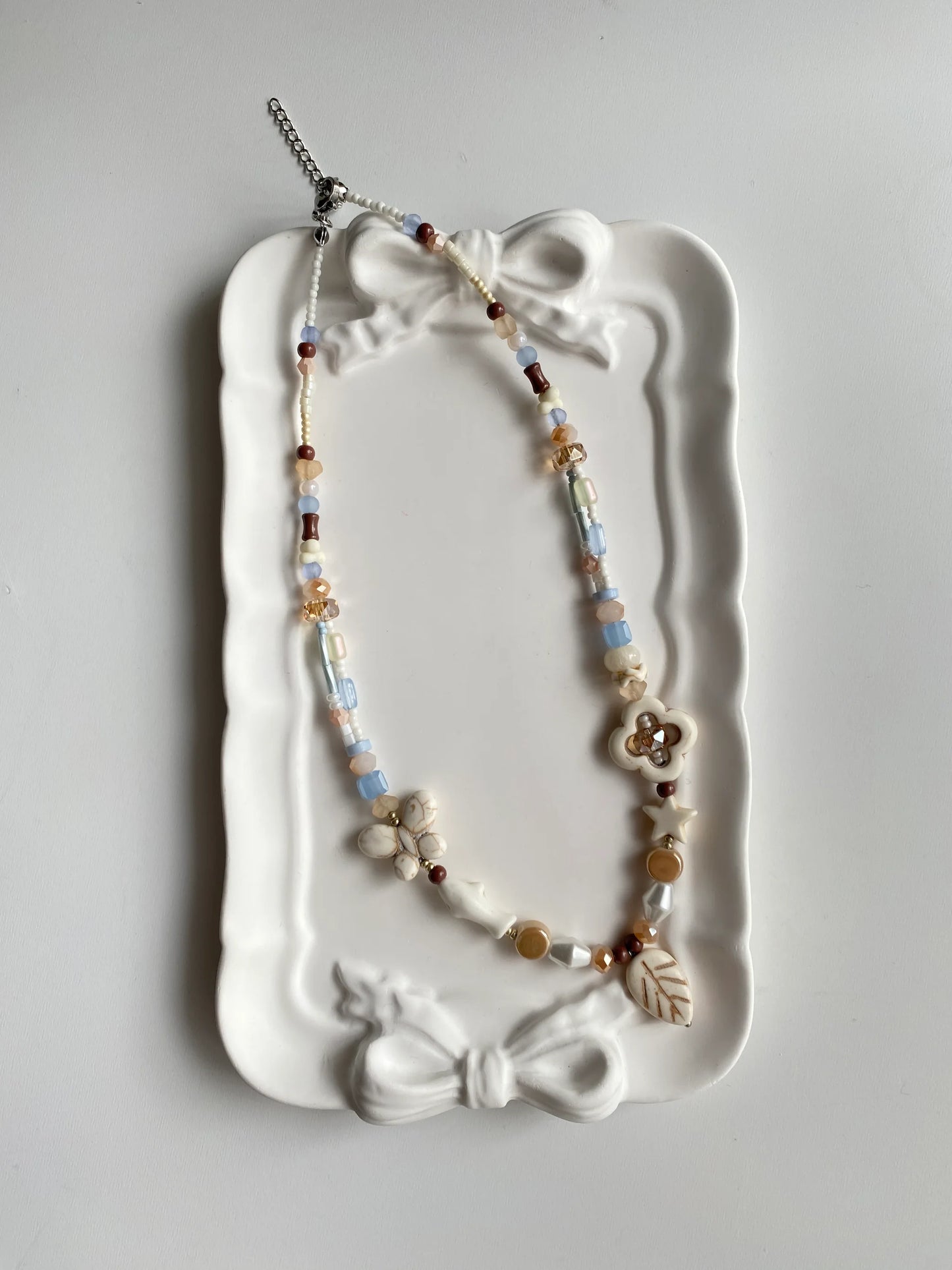 HuibenCraft 1-of-1 artisanal summer necklace with light blue and coffee-colored beads in a white ceramic dish, reflecting a relaxed holiday vibe.