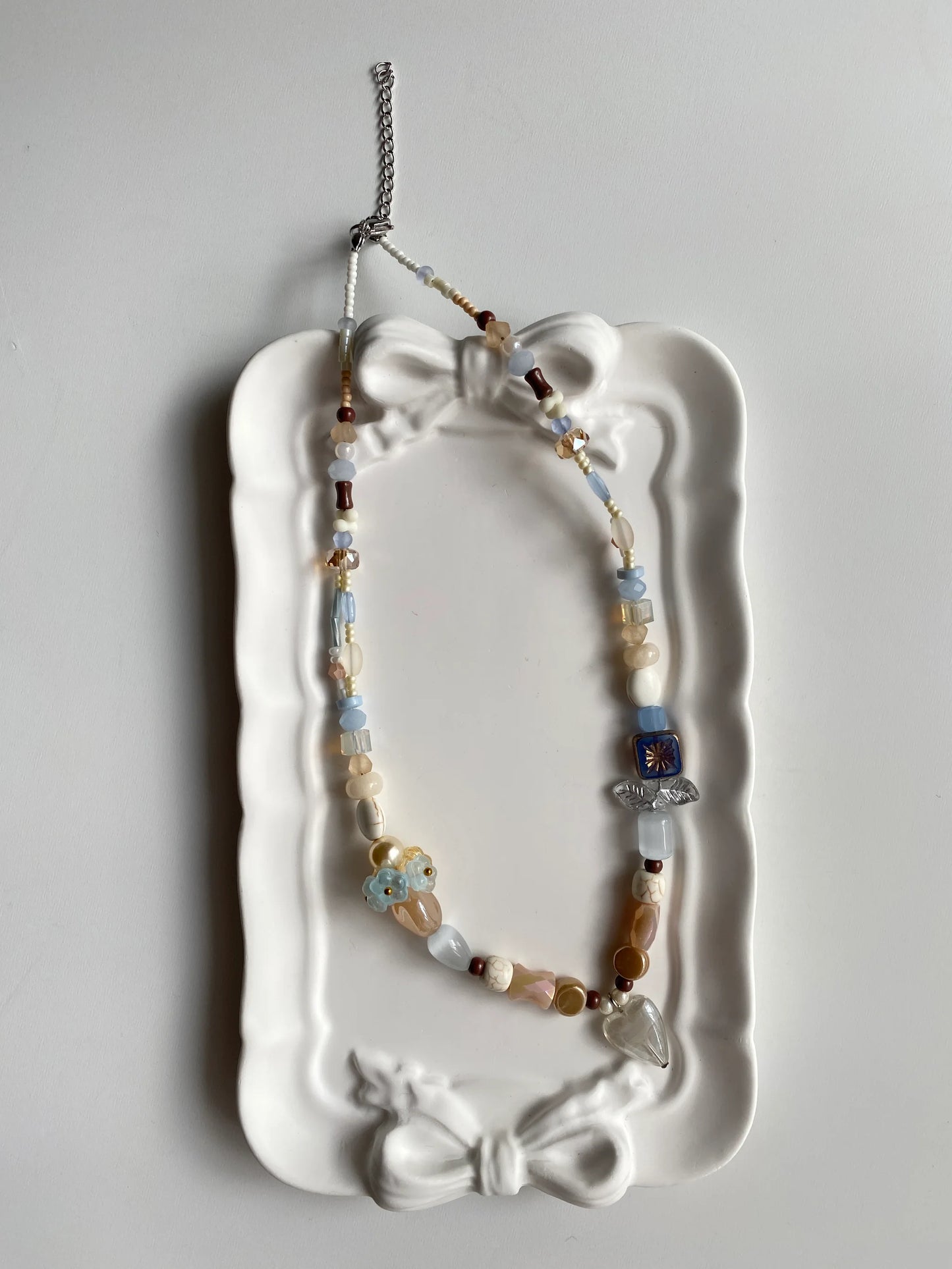 HuibenCraft 1-of-1 ethereal beaded necklace with a grey heart pendant and shimmering light-colored beads in a white ceramic dish.
