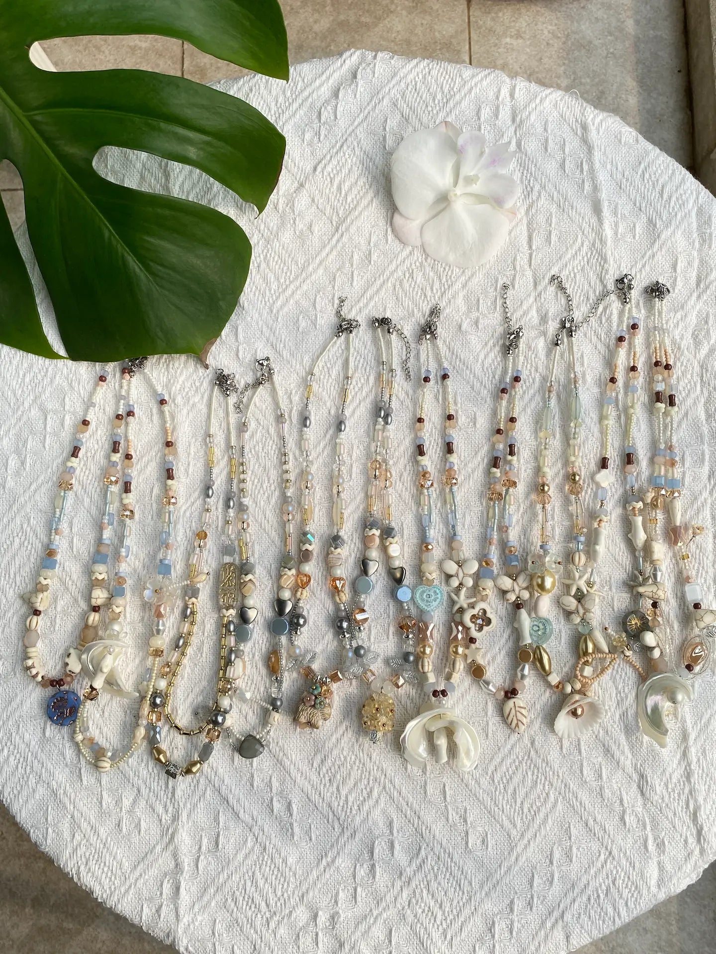 A flat lay of 10 HuibenCraft 1-of-1 coastal resort-style necklaces in sky blue and white, featuring shells and fish charms, styled with sunlight and greenery.