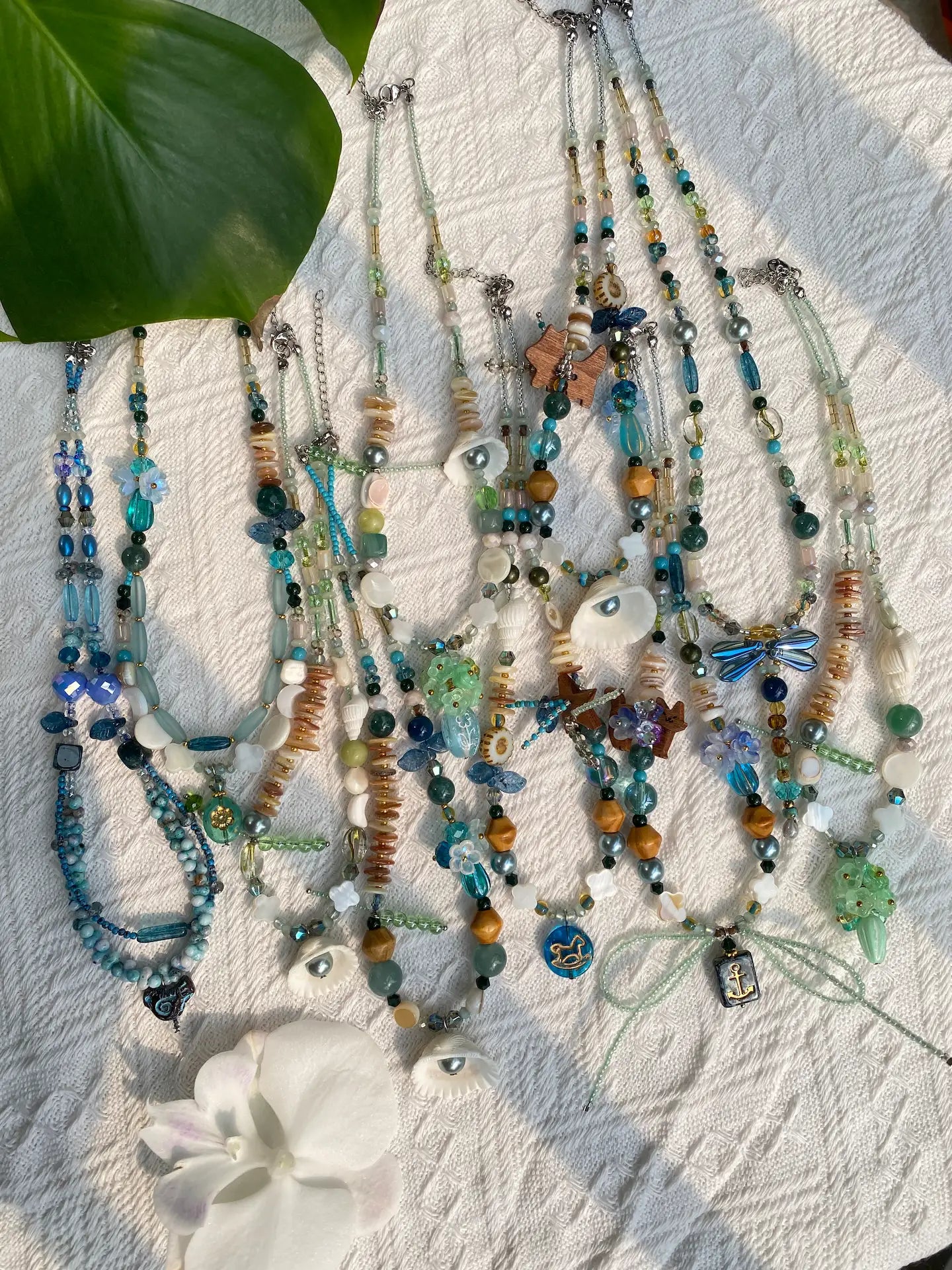 A flat lay of 10 HuibenCraft 1-of-1 ocean-themed necklaces with 3D floral beads and natural shells, captured in natural sunlight with monstera leaf shadows.