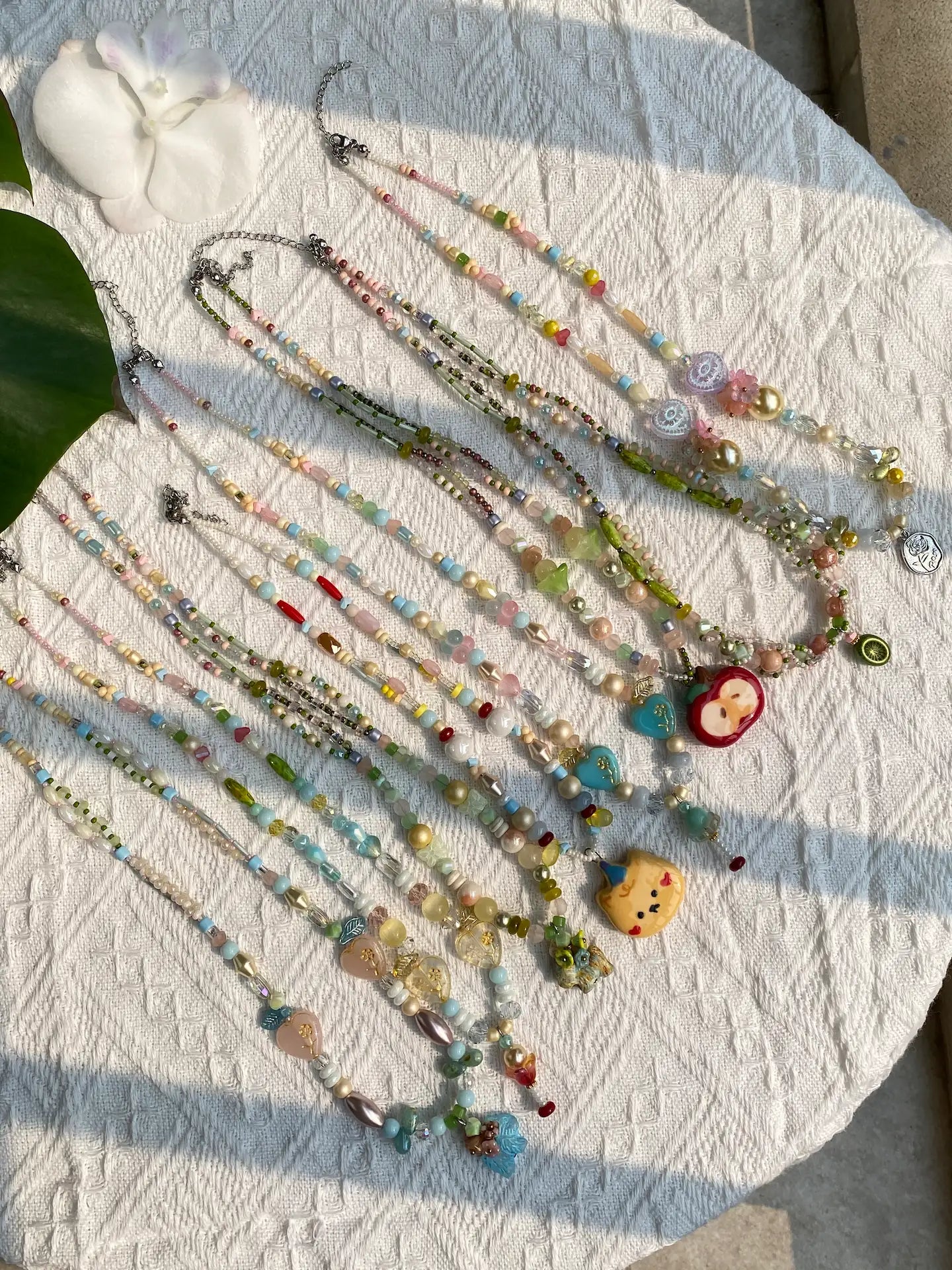 A vibrant collection of 10 HuibenCraft 1-of-1 whimsical beaded necklaces featuring ceramic bears and apples, styled on a table with soft sunlight and greenery.