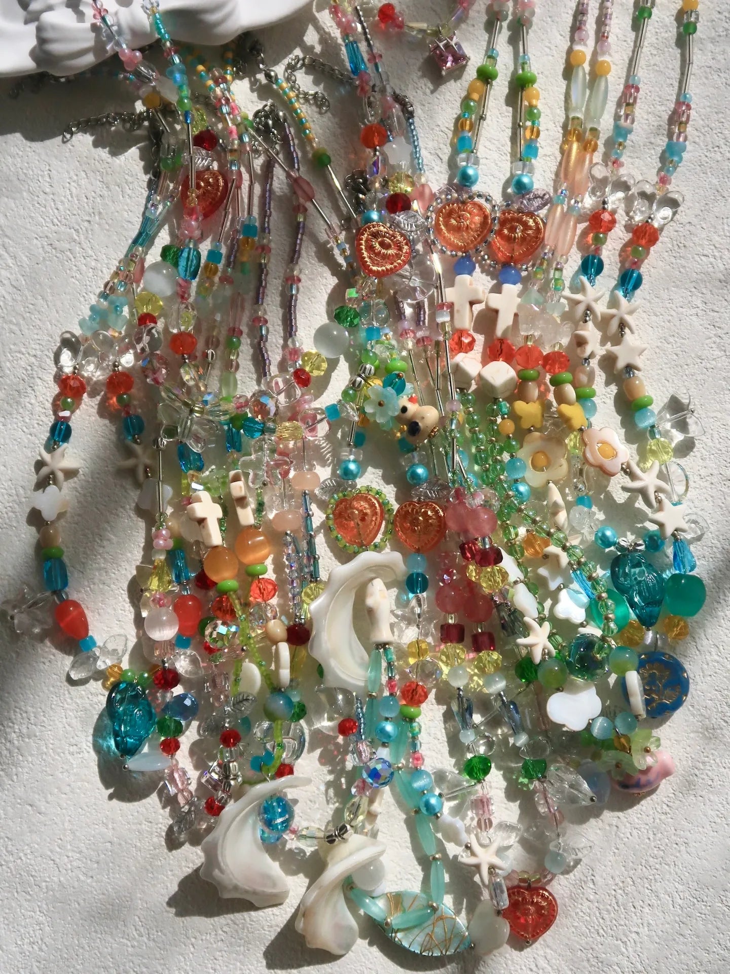 A vibrant showcase of 10 HuibenCraft 1-of-1 necklaces with Trochus shells, ceramic ducks, and colorful Czech glass shimmering on a white table in natural sunlight.