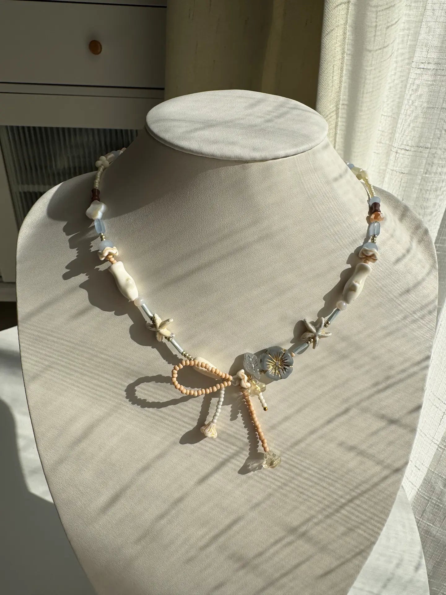 A 1-of-1 asymmetrical beaded necklace with a handmade seed bead bow and a grey gold-rimmed Czech glass flower on a jewelry bust.