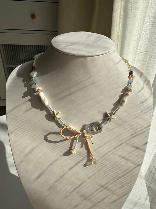 A 1-of-1 asymmetrical beaded necklace with a handmade seed bead bow and a grey gold-rimmed Czech glass flower on a jewelry bust.