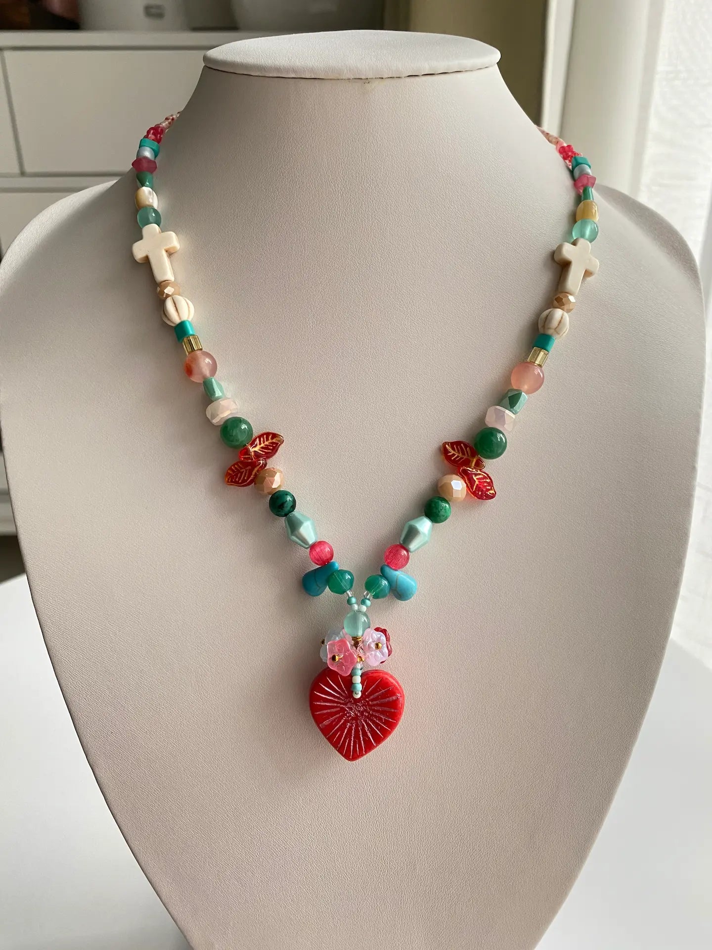 A 1-of-1 artisanal necklace featuring a large red Czech glass heart with 3D beaded flowers on a jewelry bust, pine blue and red tones.