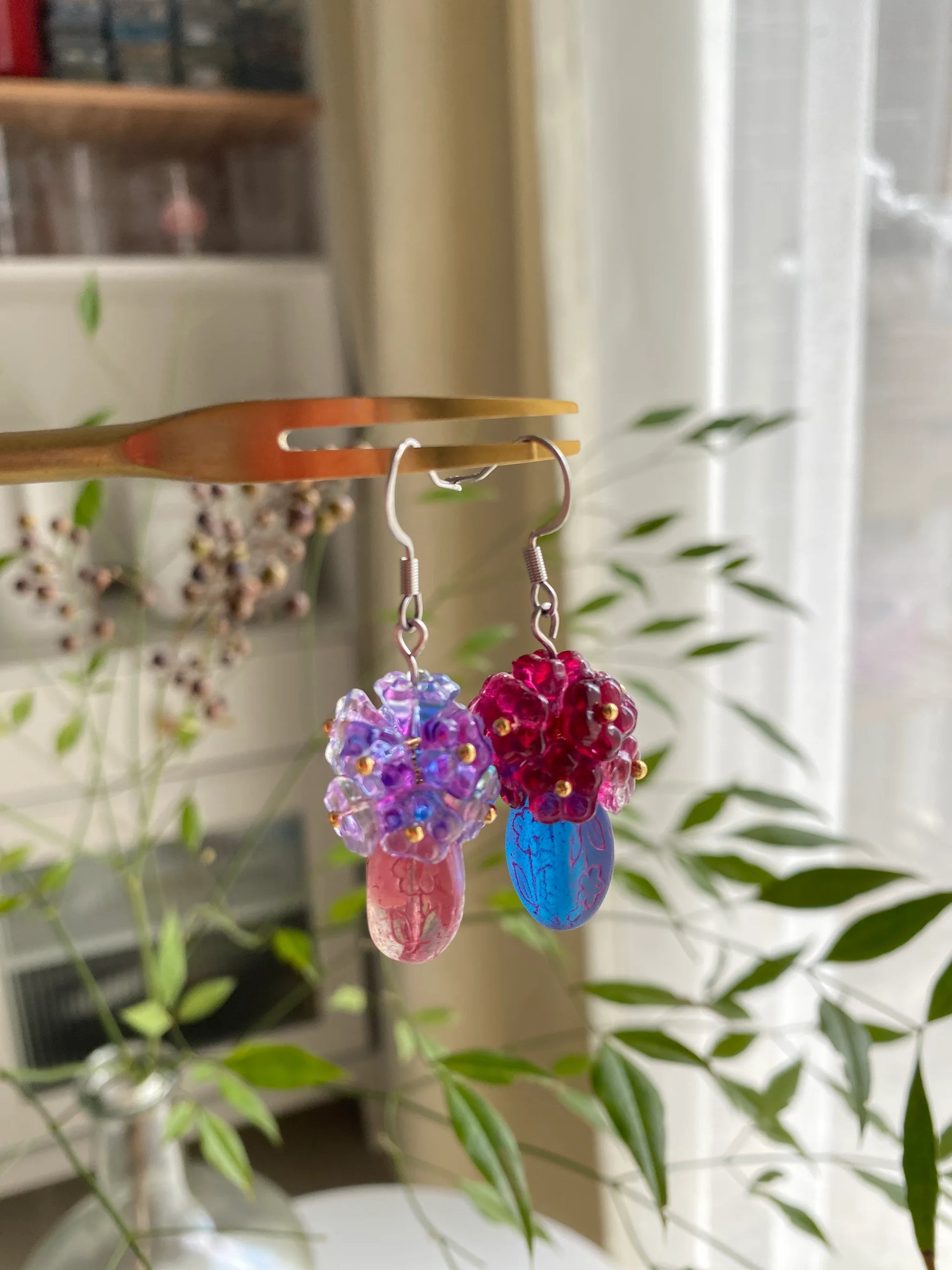 A 1-of-1 pair of handcrafted blue and purple beaded bouquet earrings on a fork, featuring a Czech glass vase with green plant background.