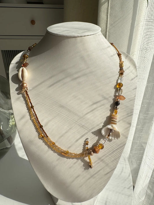A 1-of-1 double-layered bohemian long necklace in khaki and coffee tones with shell and wooden bead accents on a jewelry bust.