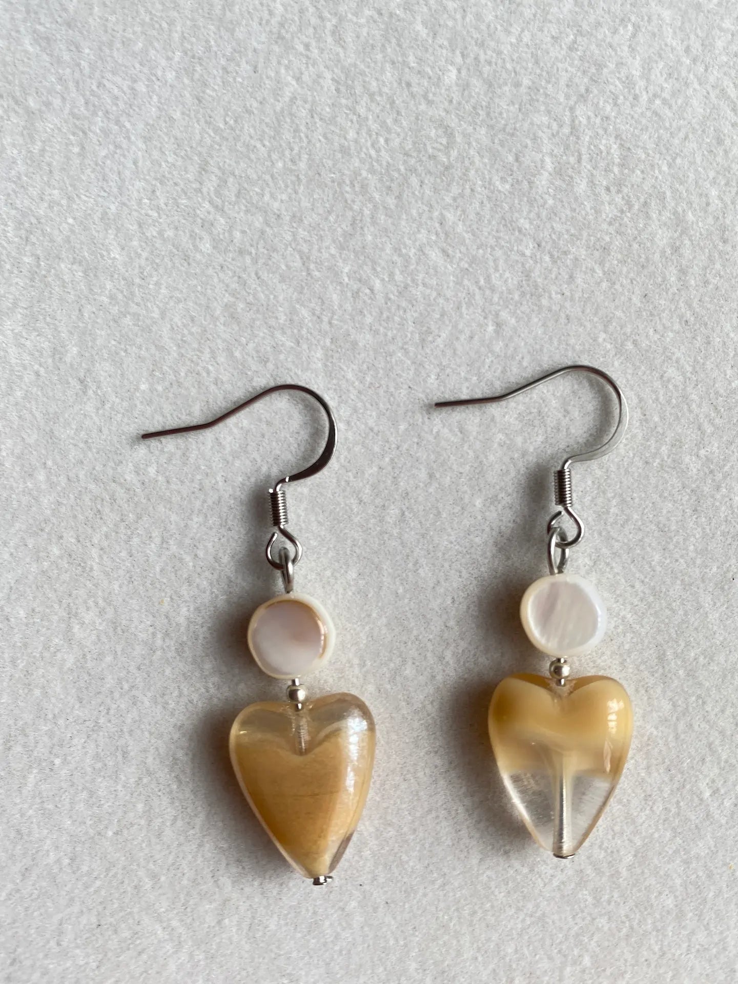 A 1-of-1 pair of cute latte-colored Czech glass heart earrings with natural round shell charms on a white tablecloth.