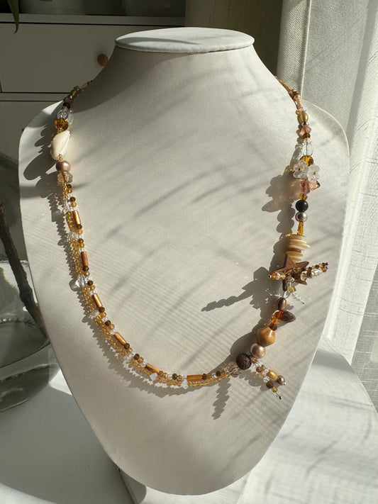 A 1-of-1 double-layered beaded necklace in khaki and brown tones with shell and coconut star charms, displayed on a jewelry bust.