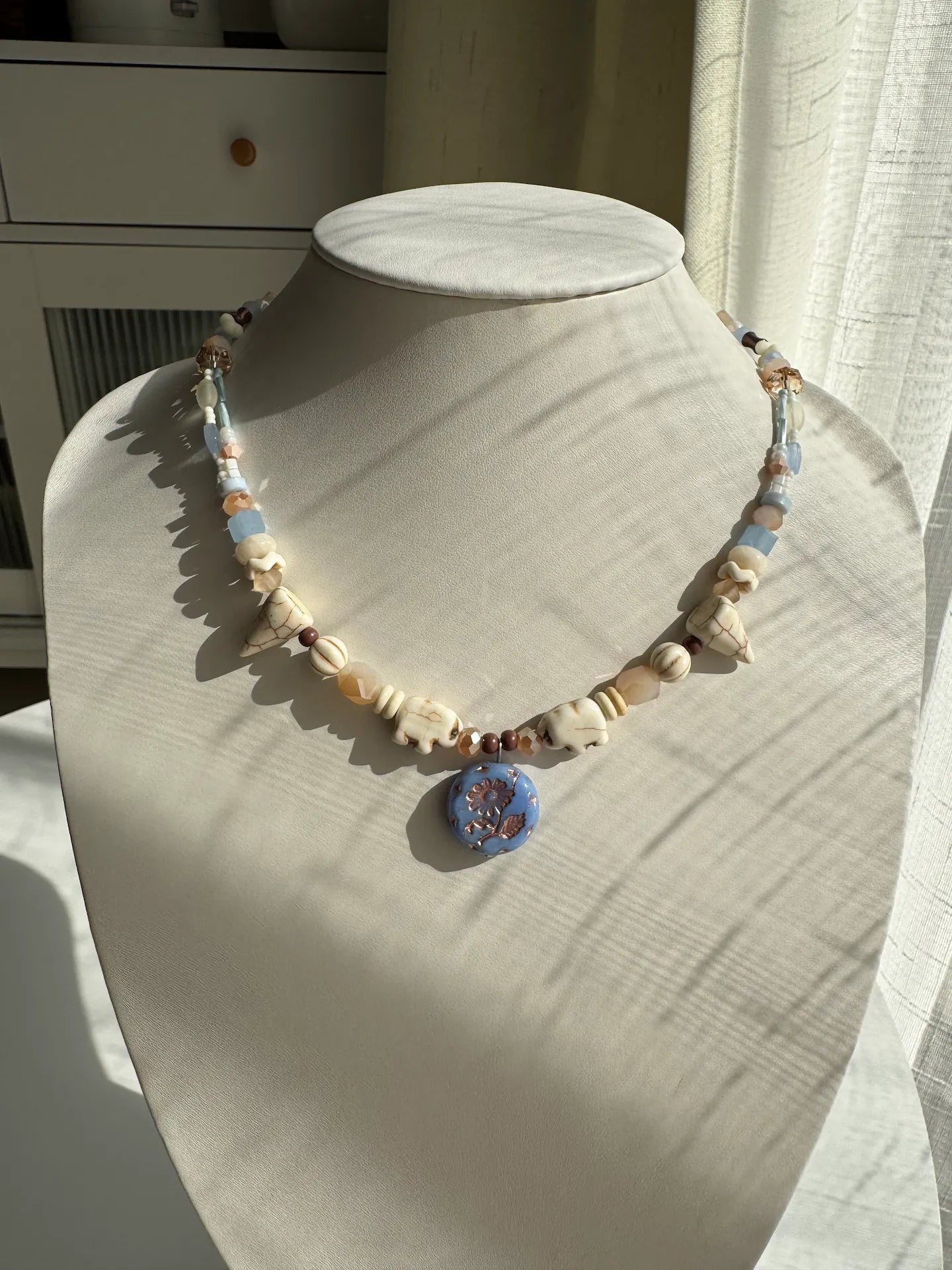 A 1-of-1 playful exotic-style necklace with white pine stone elephants and a sky blue gold-rimmed Marguerite glass pendant on a jewelry bust.