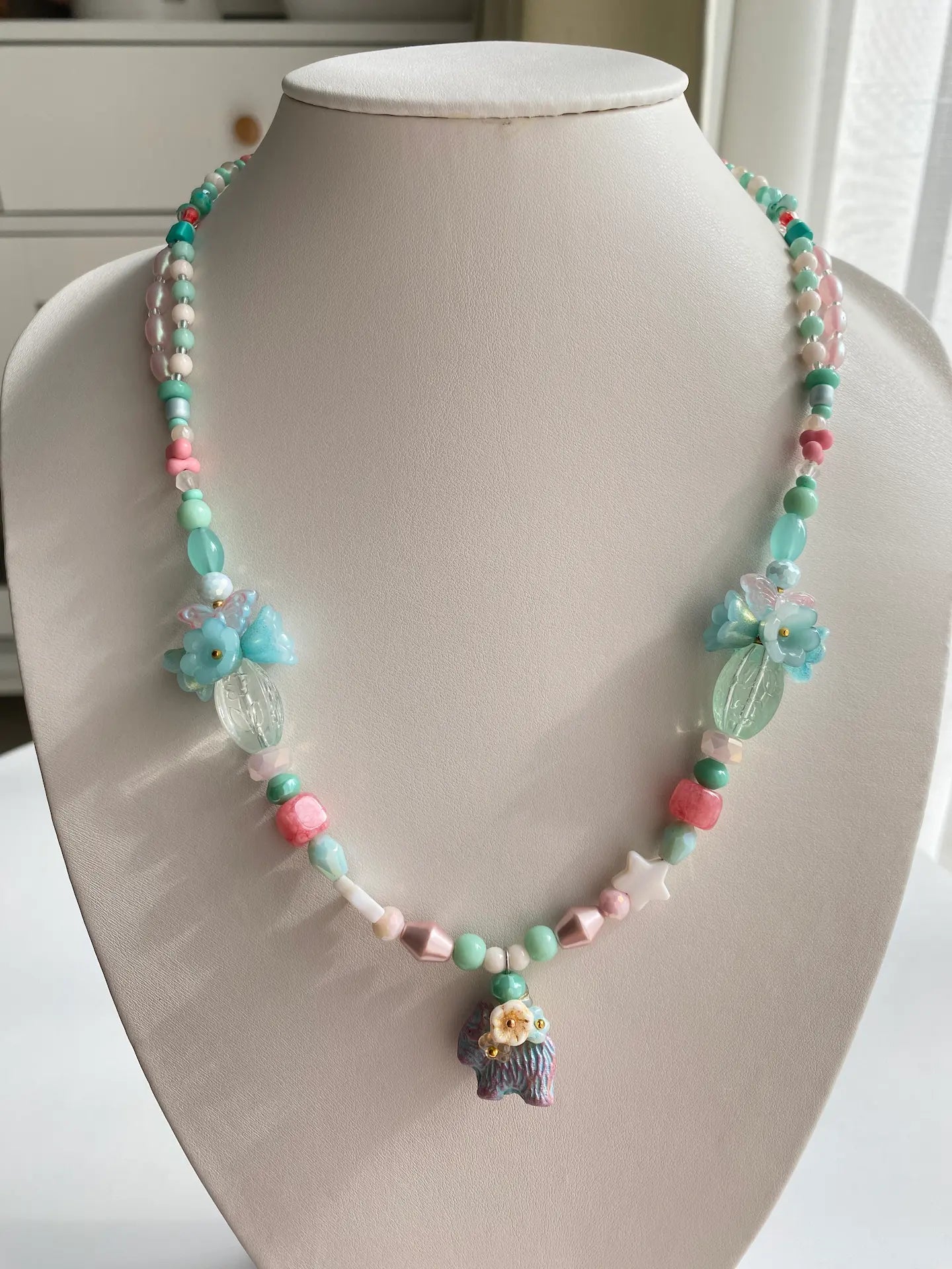 A 1-of-1 whimsical layered necklace in blue and pink with a Czech glass dog carrying a 3D beaded flower bouquet, displayed on a jewelry bust.