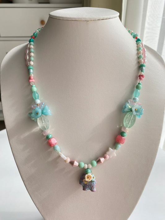 A 1-of-1 whimsical layered necklace in blue and pink with a Czech glass dog carrying a 3D beaded flower bouquet, displayed on a jewelry bust.