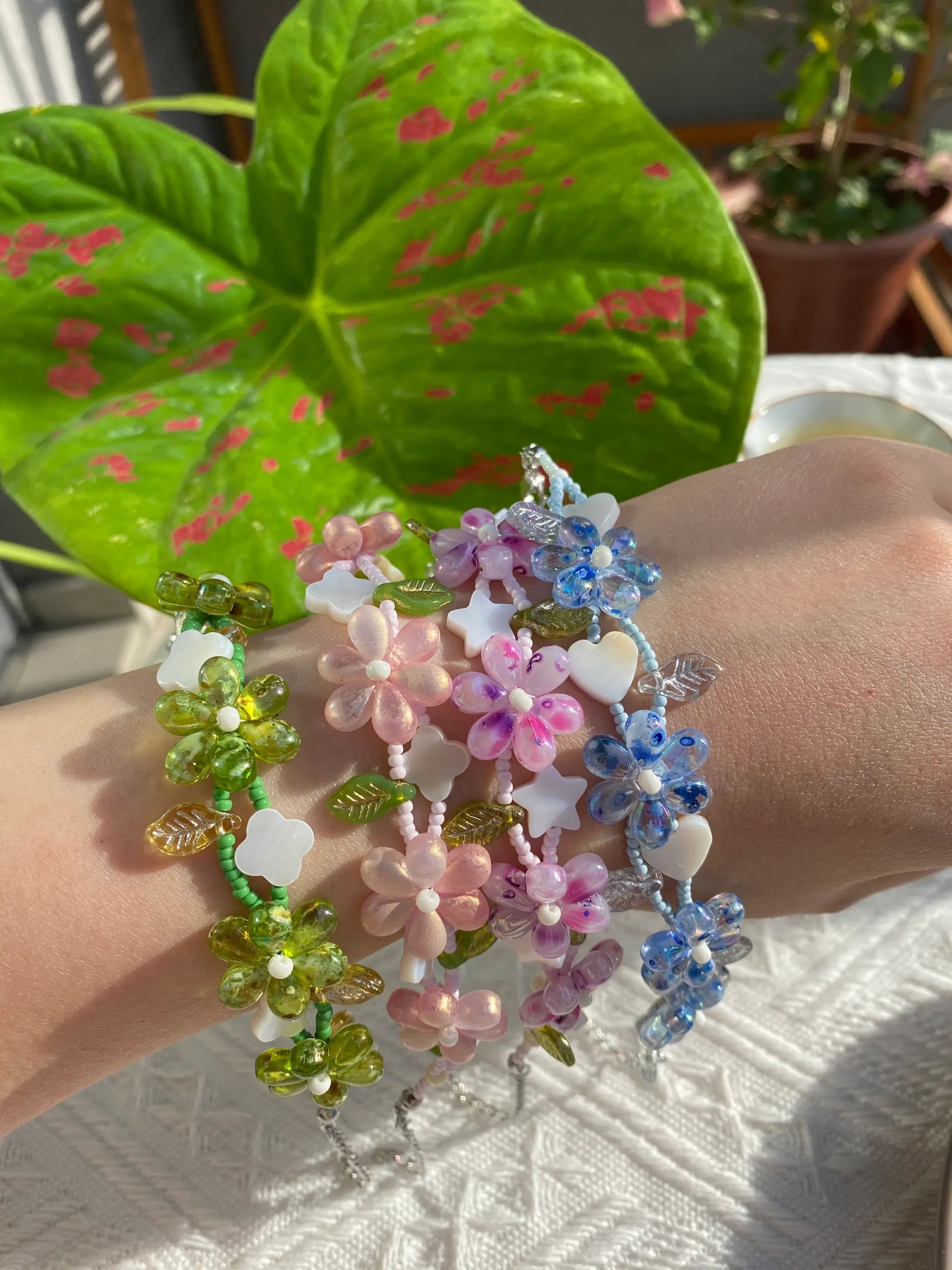 Four HuibenCraft 1-of-1 floral seed bead bracelets with unique shell charms worn on a wrist in sunny green surroundings.