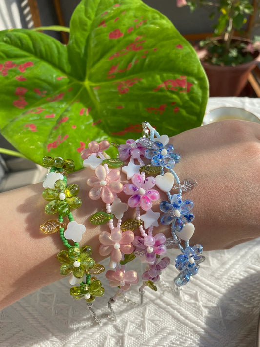 Four HuibenCraft 1-of-1 floral seed bead bracelets with unique shell charms worn on a wrist in sunny green surroundings.