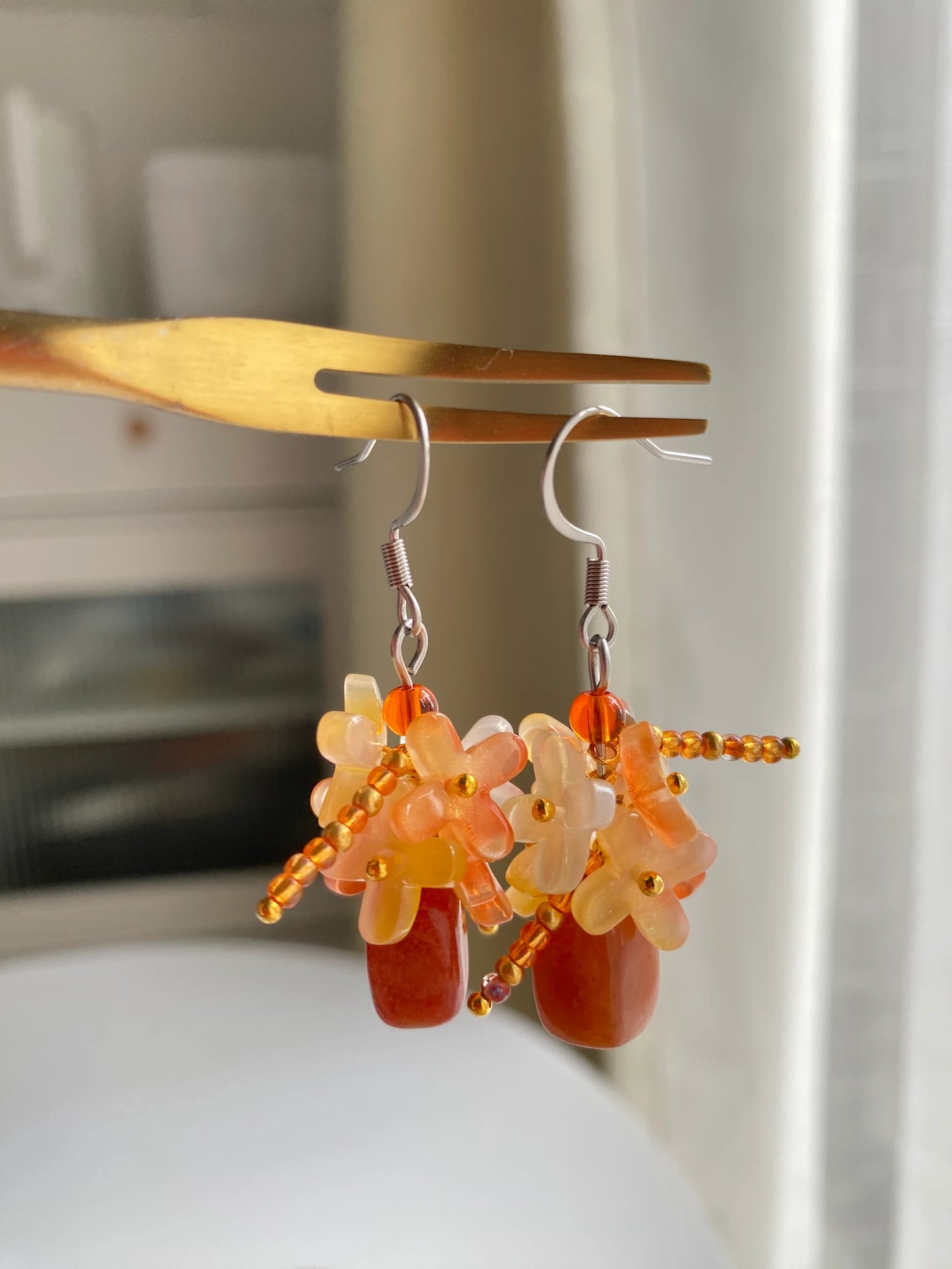 A 1-of-1 pair of majestic golden and orange earrings featuring an orange natural stone vase with a cluster of Osmanthus-shaped beads, hanging on a fork.