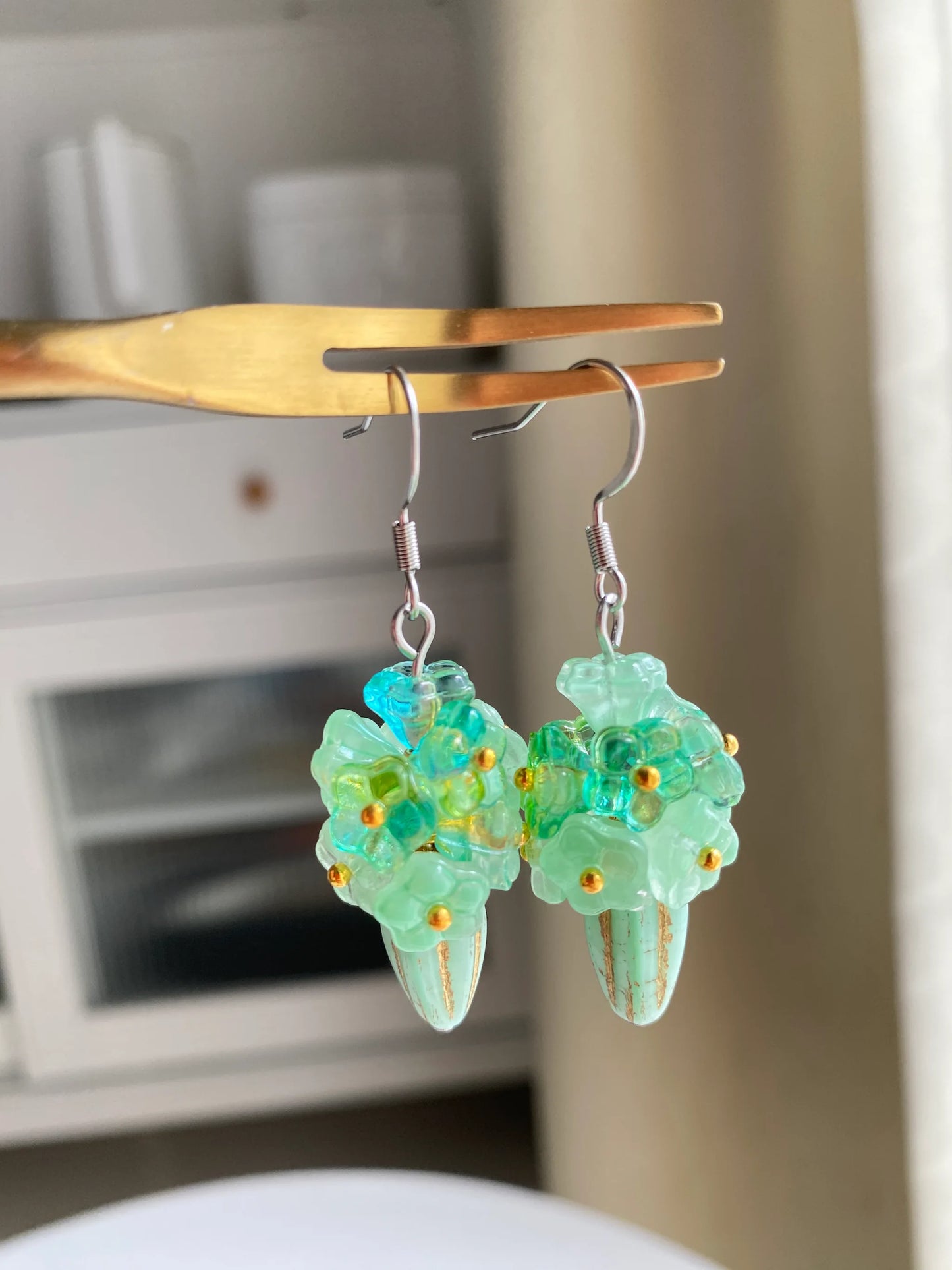 A 1-of-1 pair of lush green floral bouquet earrings featuring 11 beaded flowers in a gold-rimmed green Czech glass melon bead, hung on a fork.