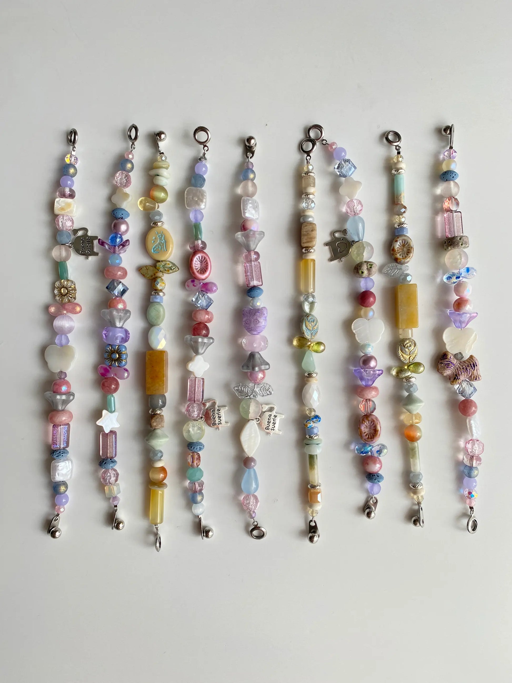 9 different handcrafted beaded bracelets in pastel colors laid out on a white table.