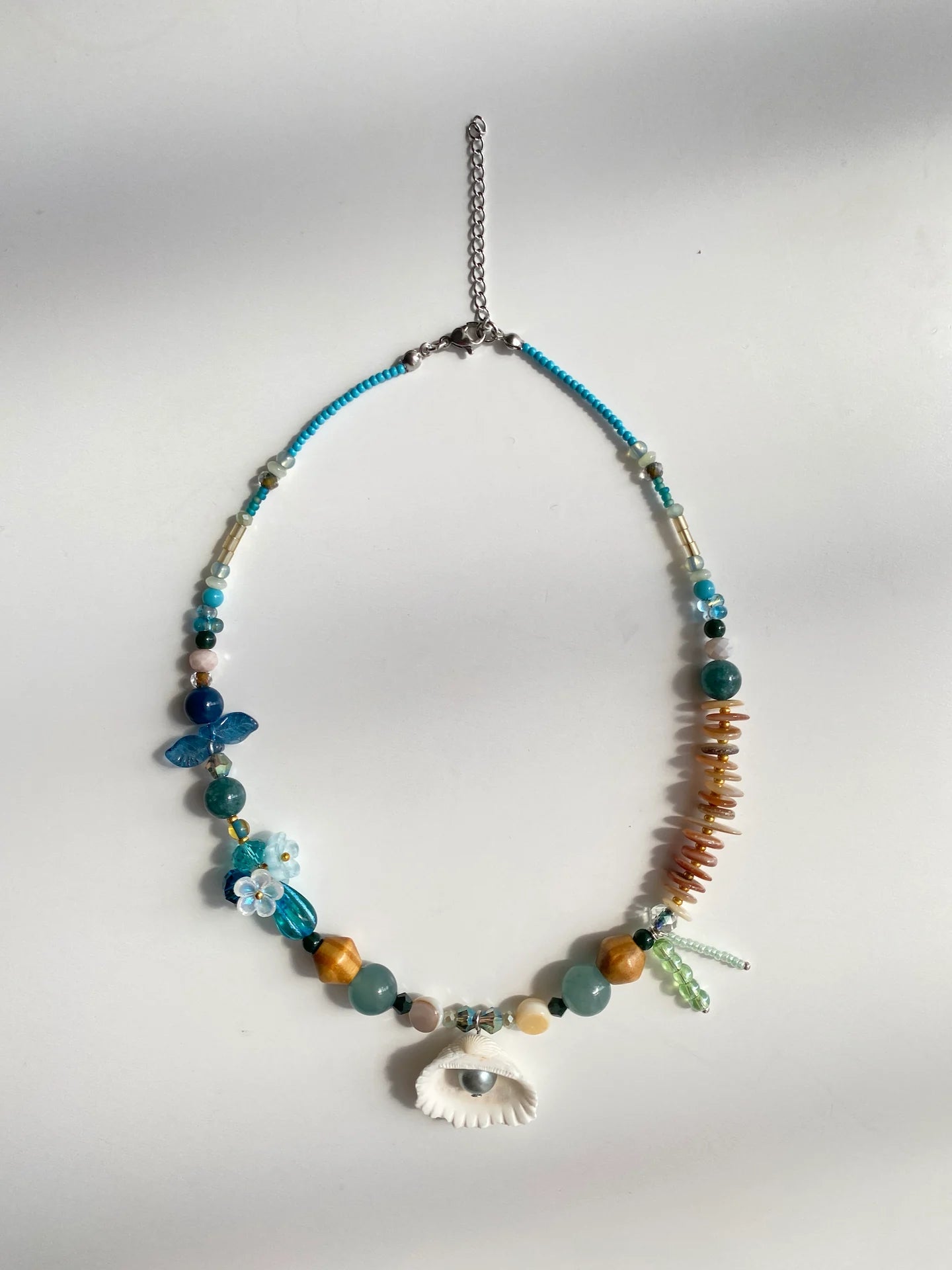 Flat lay shot on a white table of the handcrafted 1-of-1 blue necklace, clearly displaying the 3D bead flowers, shell spacers, and pendant details.