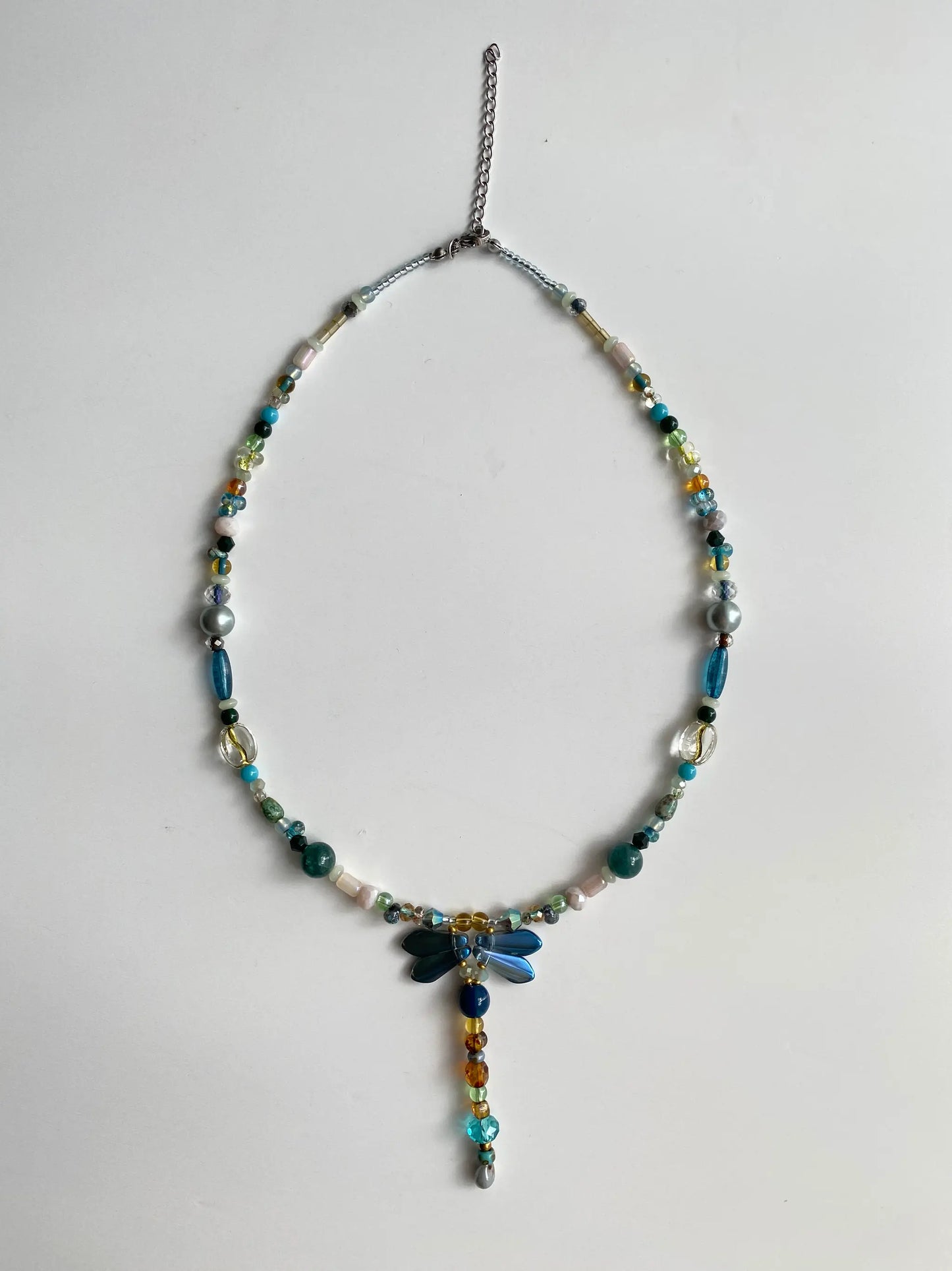 Flat lay of HuibenCraft 1-of-1 necklace on a white table, showing dragonfly details, blue stones, and specialty beads.