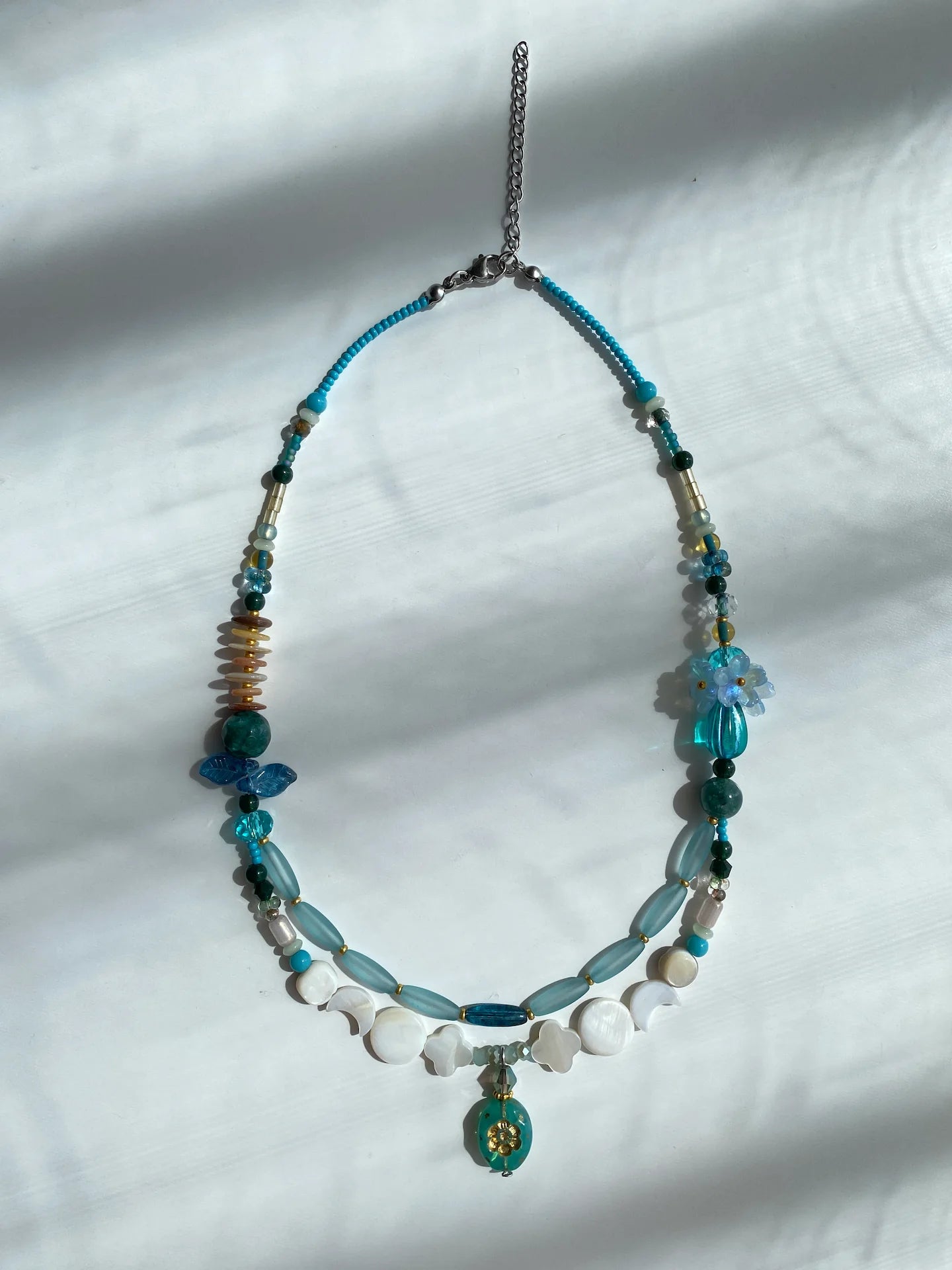 Flat lay of HuibenCraft 1-of-1 necklace on a white table, showing intricate perfume bottle pendant and layered shell/bead details.