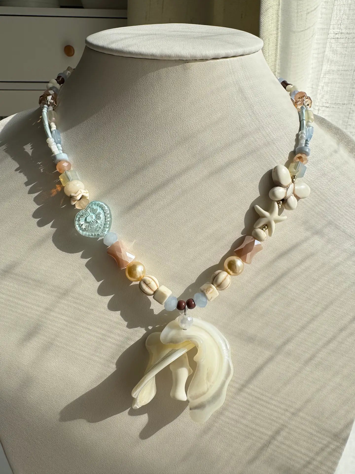 A 1-of-1 ocean vacation-style necklace with a large double trochus shell pendant and white pine star charms on a jewelry bust, blue and pink tones.
