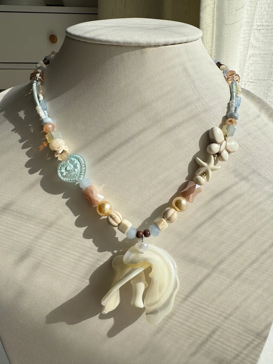 A 1-of-1 ocean vacation-style necklace with a large double trochus shell pendant and white pine star charms on a jewelry bust, blue and pink tones.