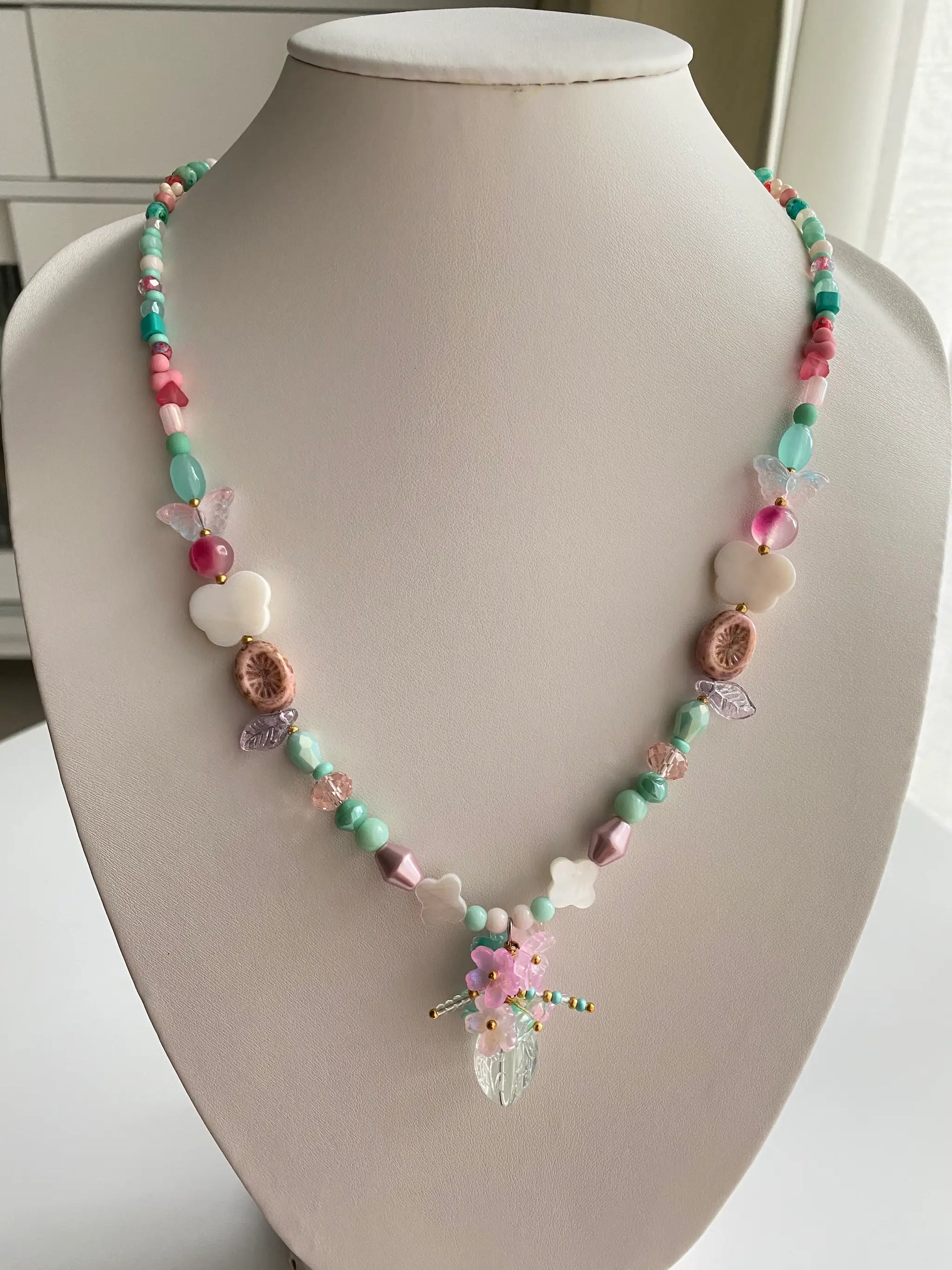 A 1-of-1 pink long beaded necklace featuring a handcrafted floral vase pendant with shimmering shell butterflies and clovers on a jewelry bust.