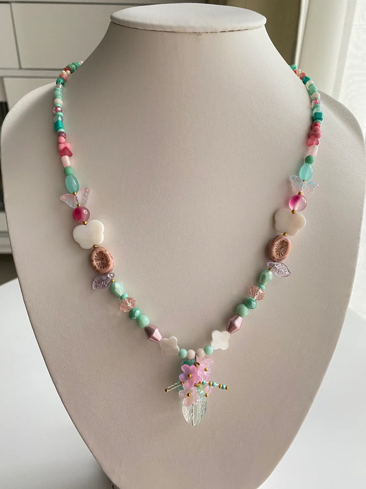 A 1-of-1 pink long beaded necklace featuring a handcrafted floral vase pendant with shimmering shell butterflies and clovers on a jewelry bust.