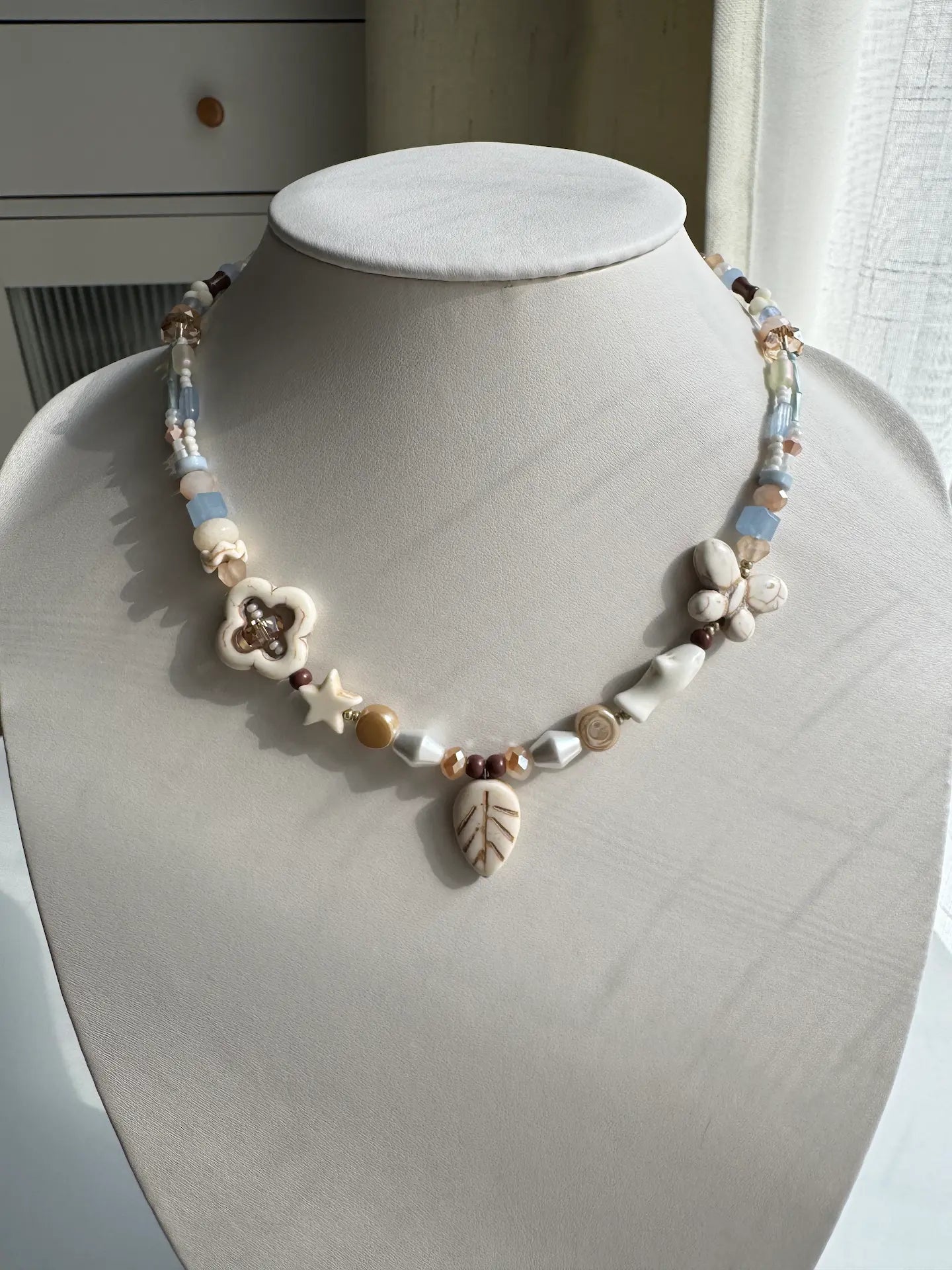 A 1-of-1 relaxed vacation-style necklace with white pine stone leaf pendant and clover charms on a jewelry bust, sky blue and white tones.
