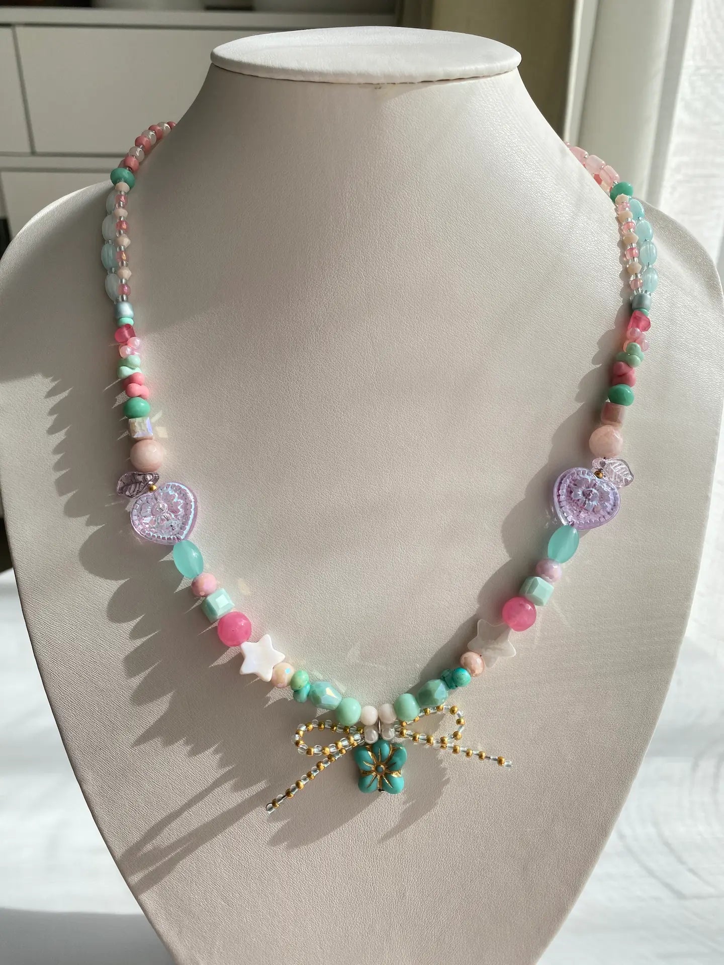 A 1-of-1 lively beaded necklace in sky blue and pink with a starfish-shaped Czech glass pendant and a handmade seed bead bow on a jewelry bust.