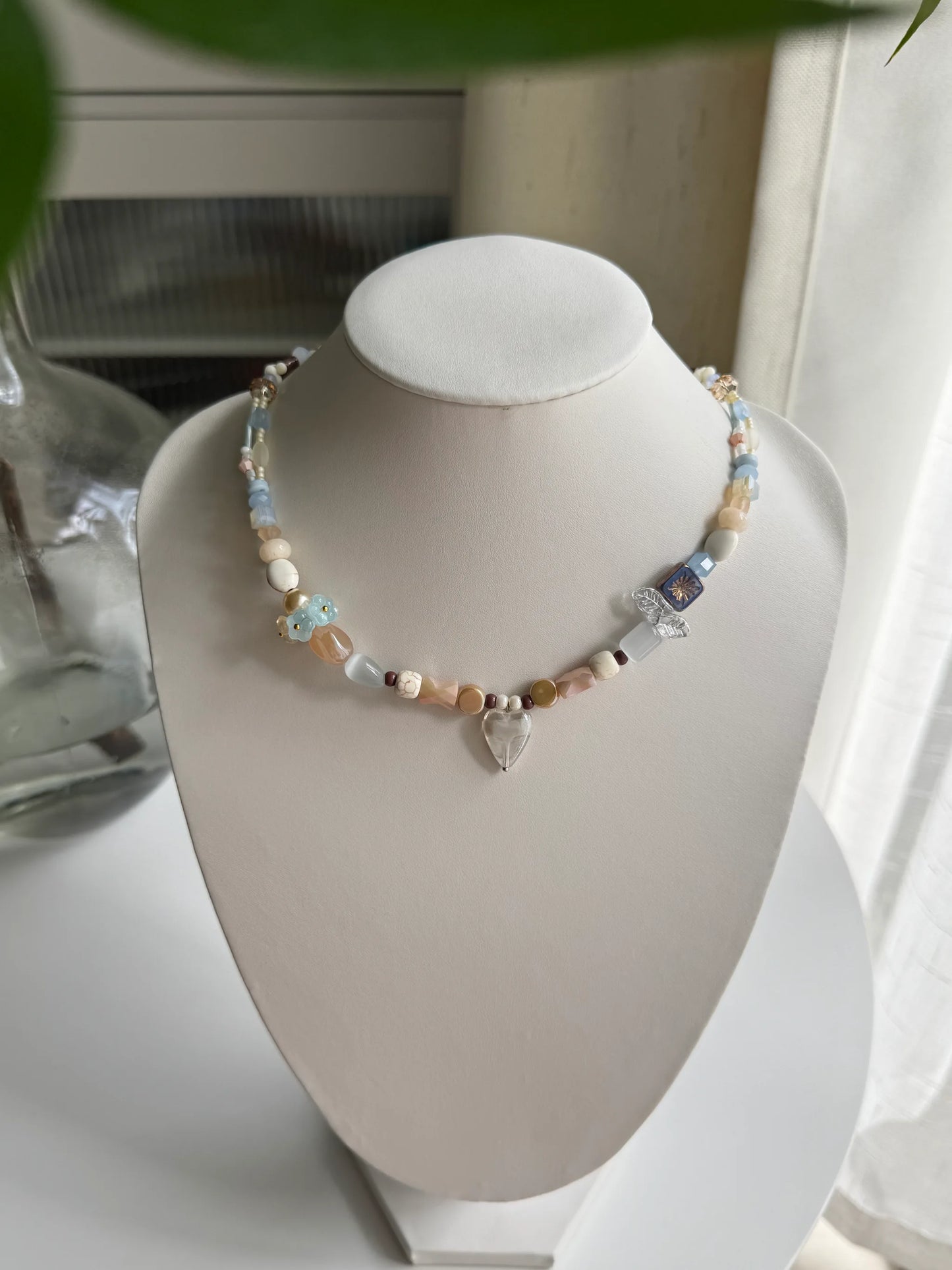 A 1-of-1 gentle vacation-style beaded necklace with a translucent grey heart pendant and fireworks-patterned glass on a jewelry bust.
