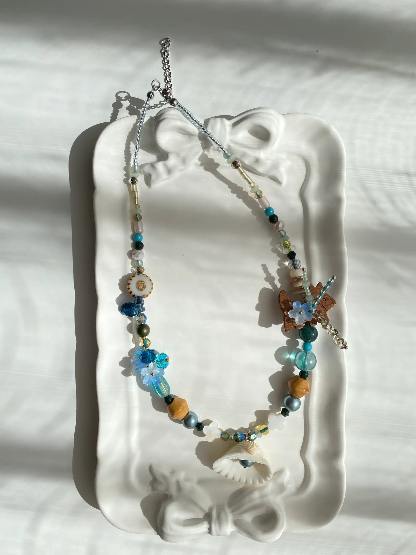 Poetic view of the 1-of-1 ocean-themed necklace with a shell pendant resting on a ceramic plate, capturing a relaxed vacation aesthetic.