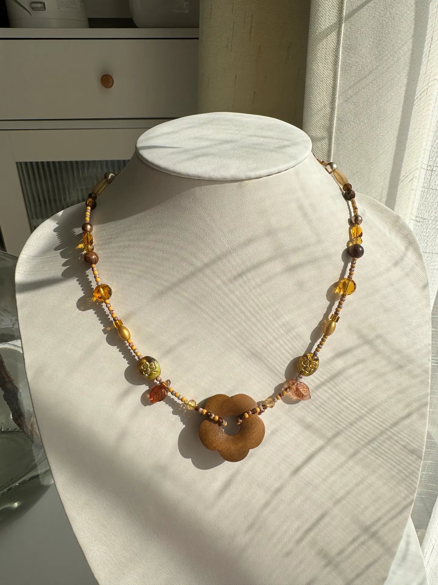 A 1-of-1 handcrafted beaded necklace in khaki and gold tones with a large wooden flower pendant on a jewelry bust.
