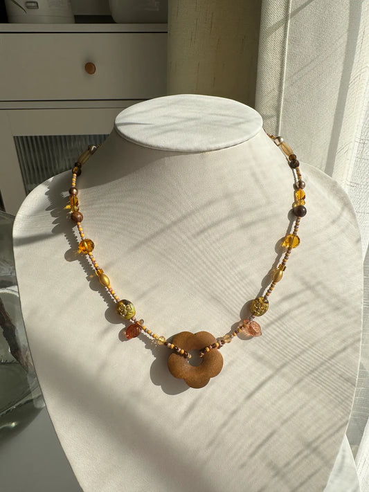 A 1-of-1 handcrafted beaded necklace in khaki and gold tones with a large wooden flower pendant on a jewelry bust.