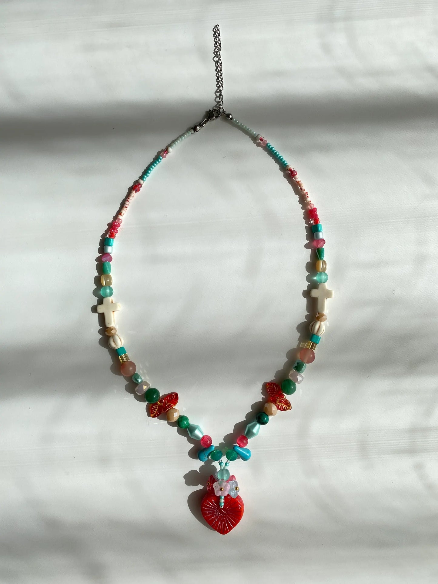 Close-up of a unique red heart pendant layered with a bouquet of colorful beads on a sunlit white desk, symbolizing "flowers in the heart."