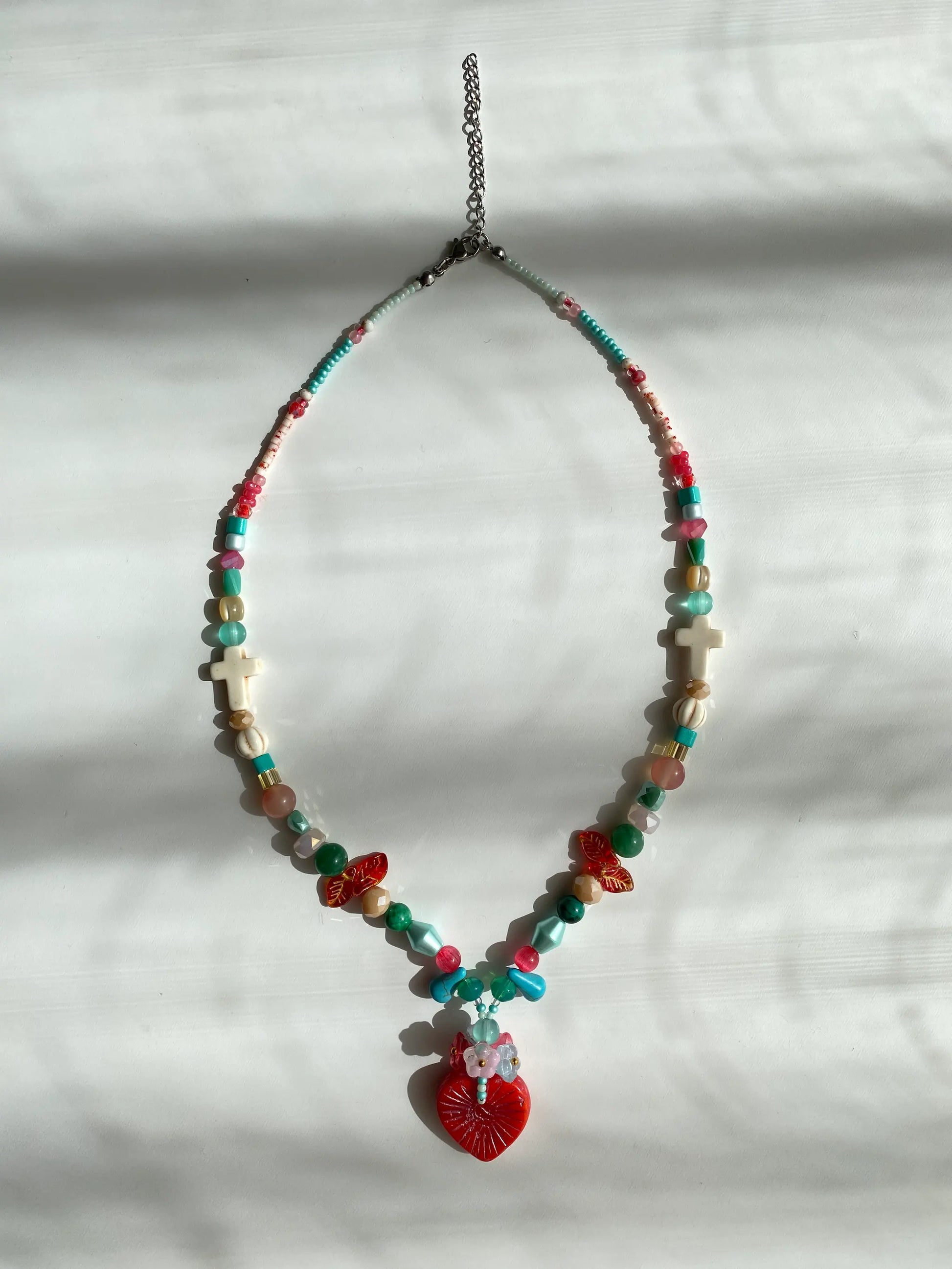 Close-up of a unique red heart pendant layered with a bouquet of colorful beads on a sunlit white desk, symbolizing "flowers in the heart."