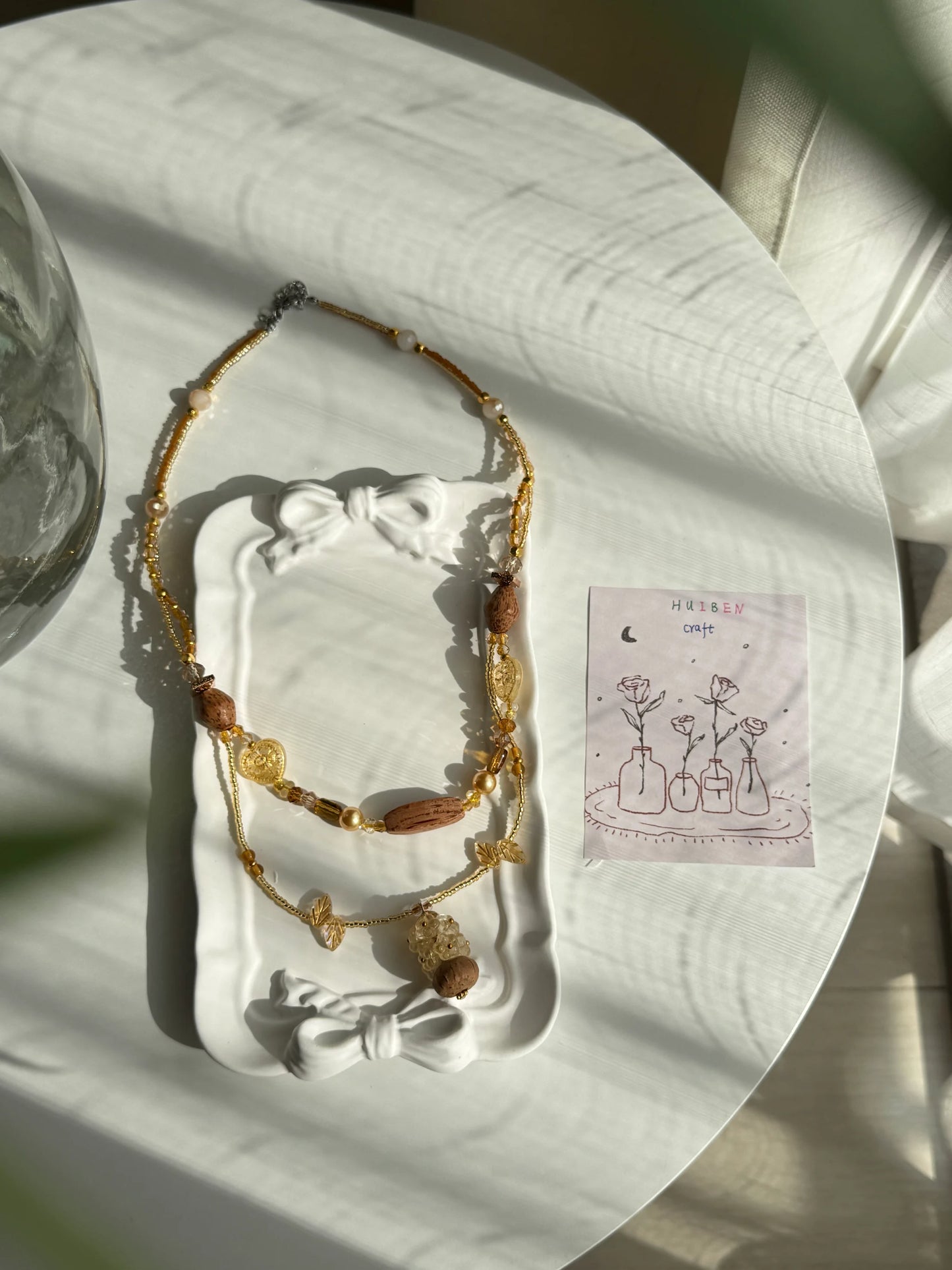 HuibenCraft 1-of-1 shimmering double-layered long necklace with heart-shaped glass beads and a floral wooden vase in a white ceramic dish.