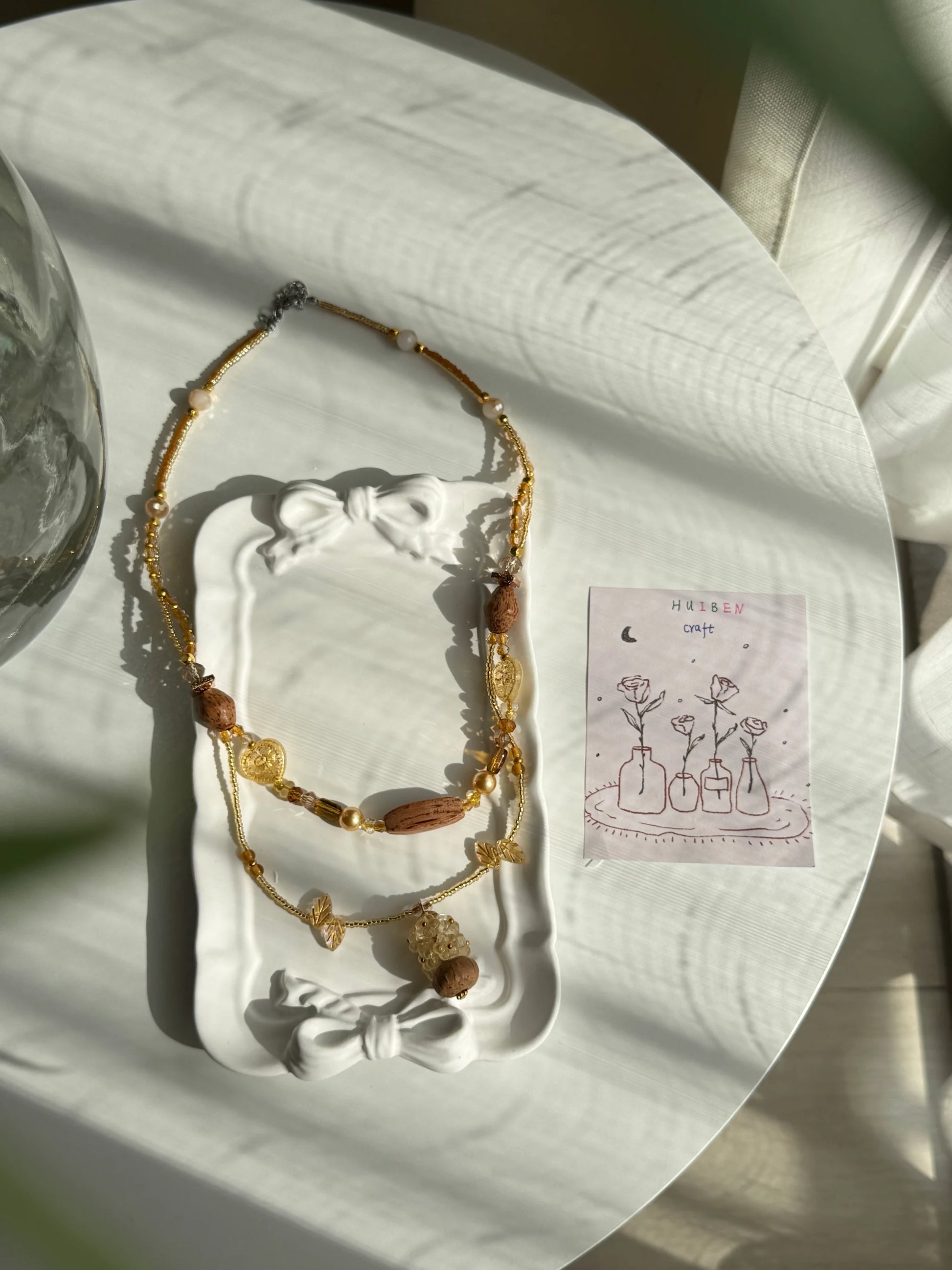 HuibenCraft 1-of-1 shimmering double-layered long necklace with heart-shaped glass beads and a floral wooden vase in a white ceramic dish.