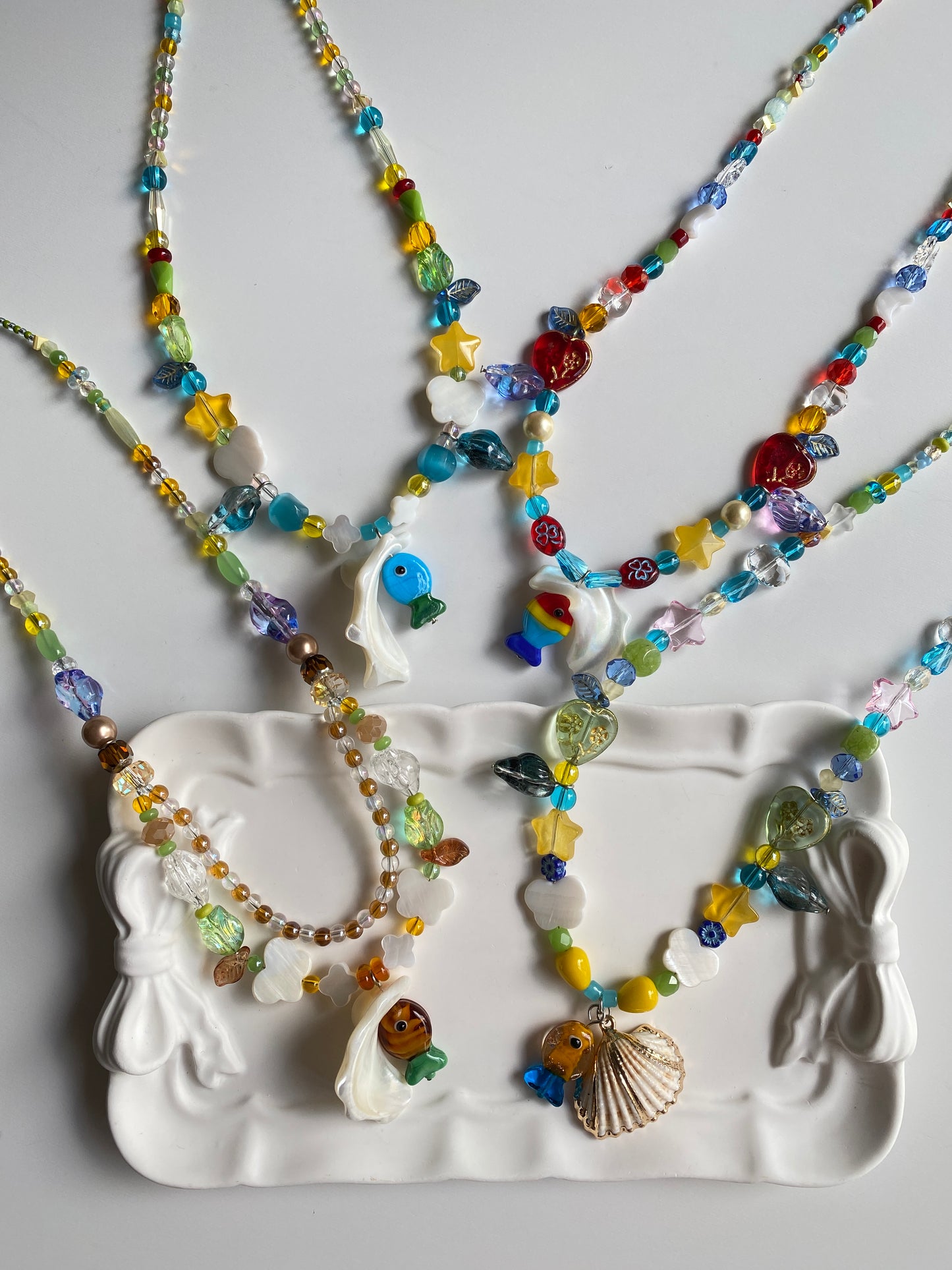 Four one-of-one handmade ocean-inspired fish necklaces displayed together on a white ceramic plate in natural sunlight, featuring colorful Czech glass fish, natural shells and coastal charms.
