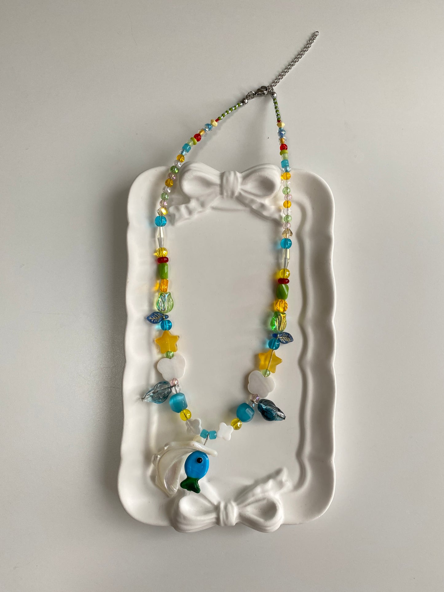 Close-up detail of a handmade ocean necklace featuring a blue-green glass fish, dark blue glass conch charm, translucent tulip bead and clover shell accents.