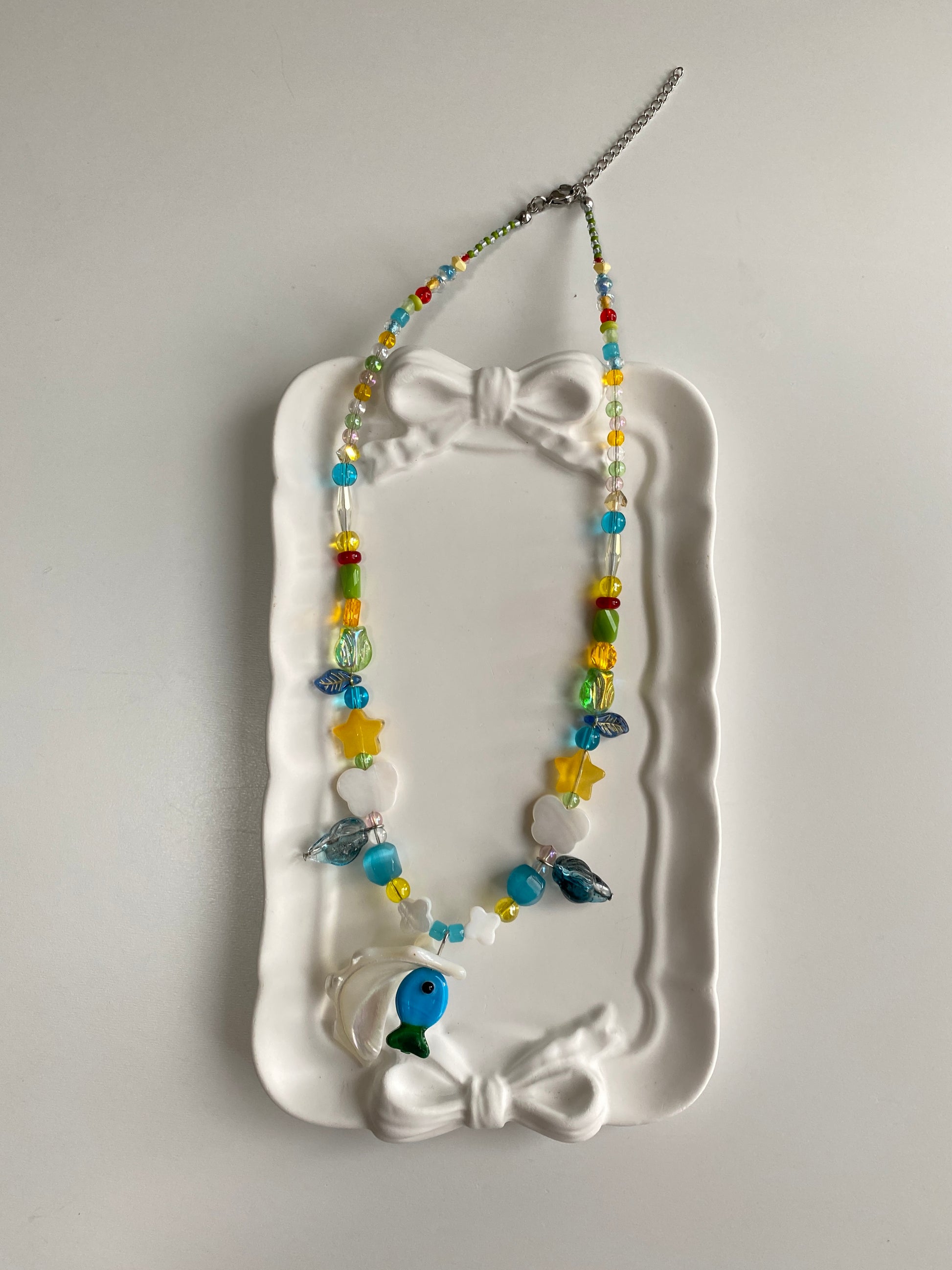 Close-up detail of a handmade ocean necklace featuring a blue-green glass fish, dark blue glass conch charm, translucent tulip bead and clover shell accents.