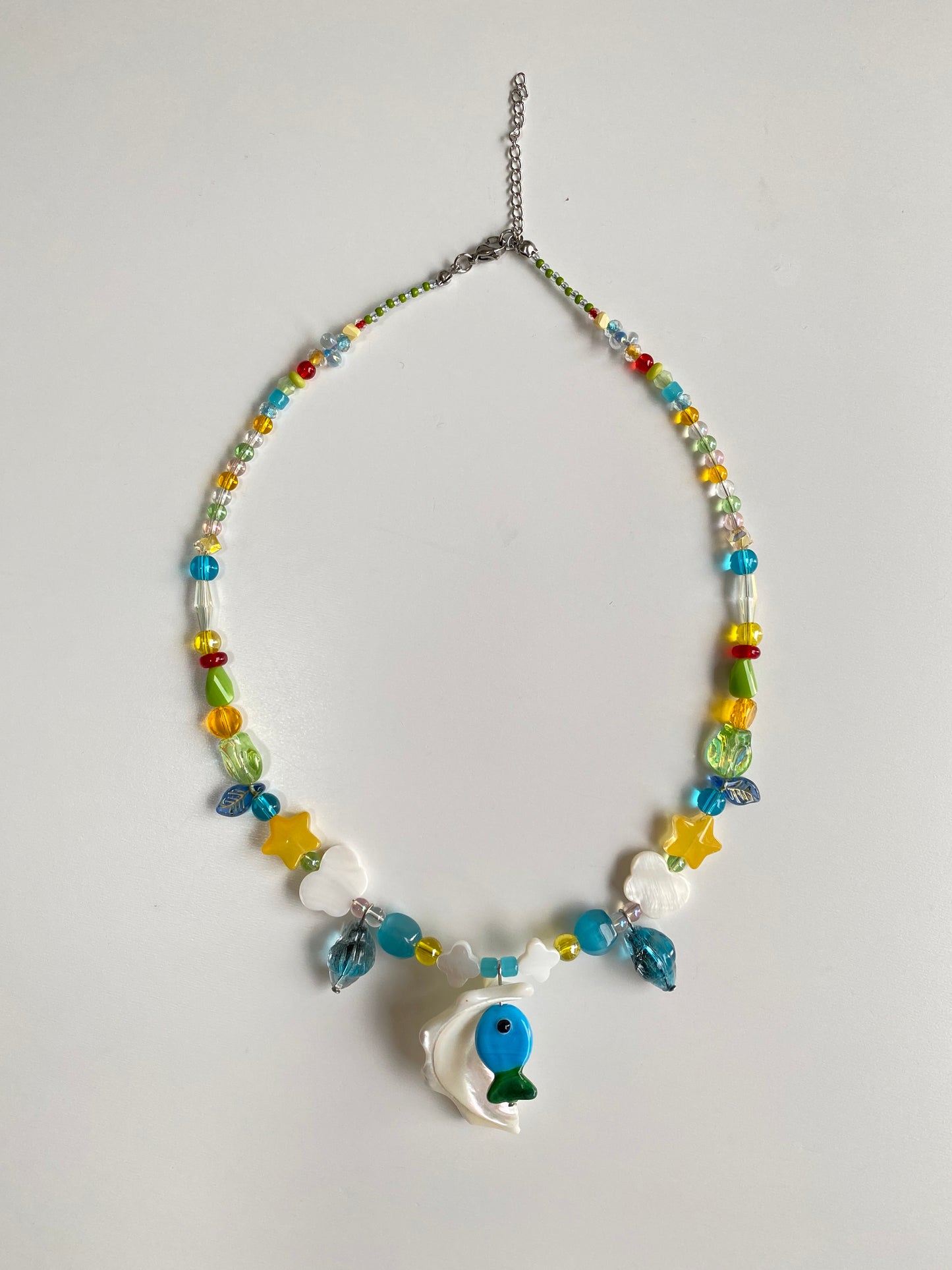 Blue-green Czech glass fish necklace displayed flat on a white background with a natural Triton shell pendant, butterfly shell charms and colorful seed beads.