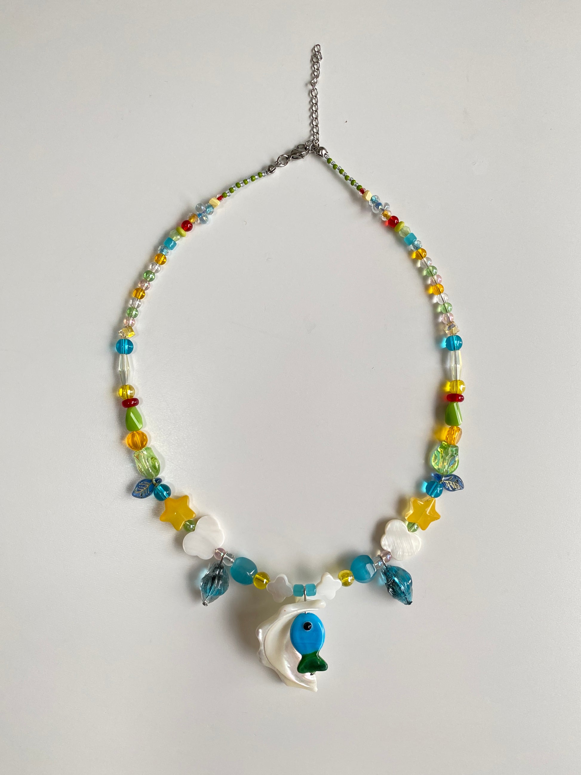 Blue-green Czech glass fish necklace displayed flat on a white background with a natural Triton shell pendant, butterfly shell charms and colorful seed beads.
