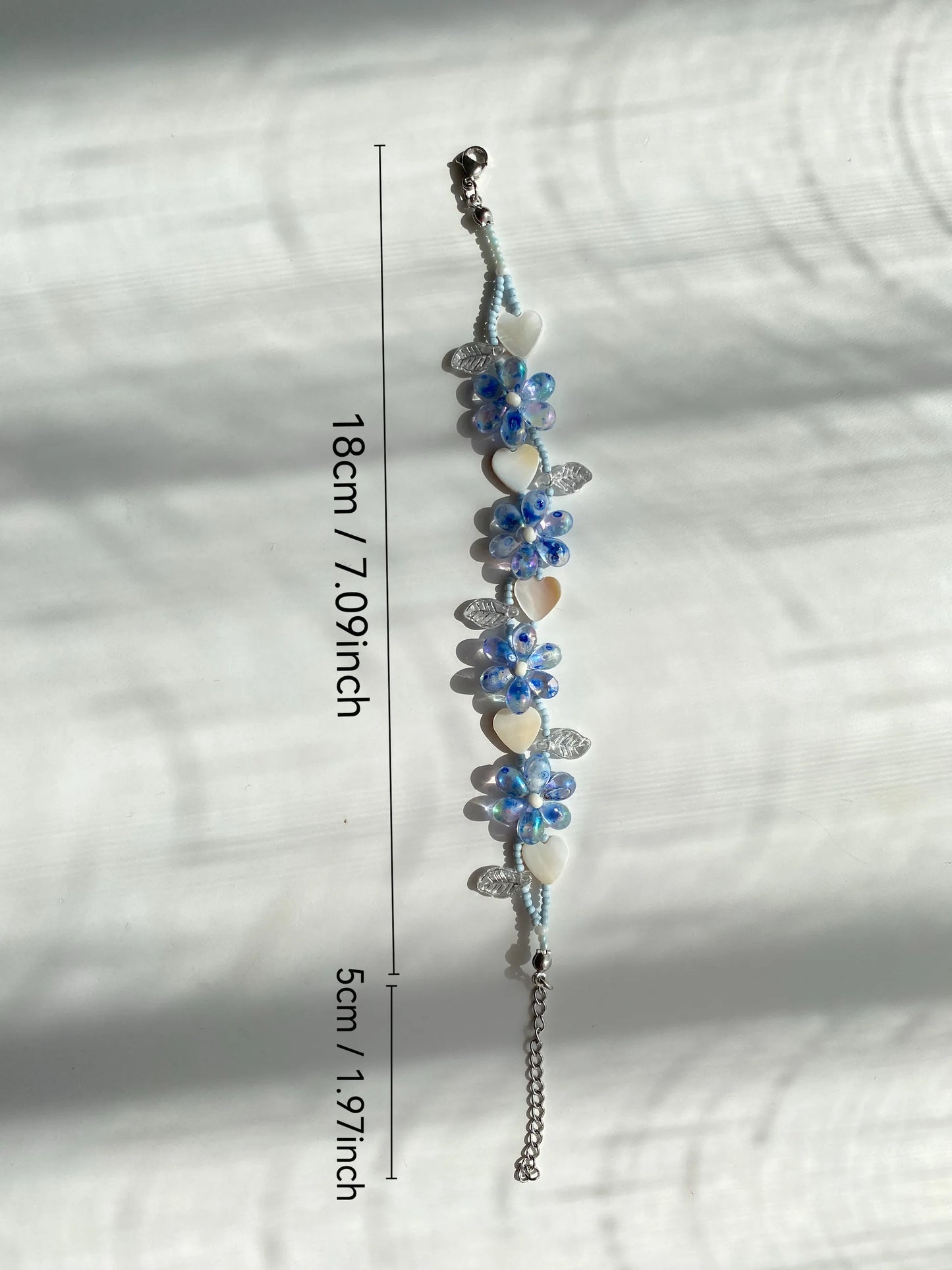 A 1-of-1 blue beaded bracelet featuring a unique waterdrop floral bloom and a heart-shaped shell charm on a sunlit white desk.