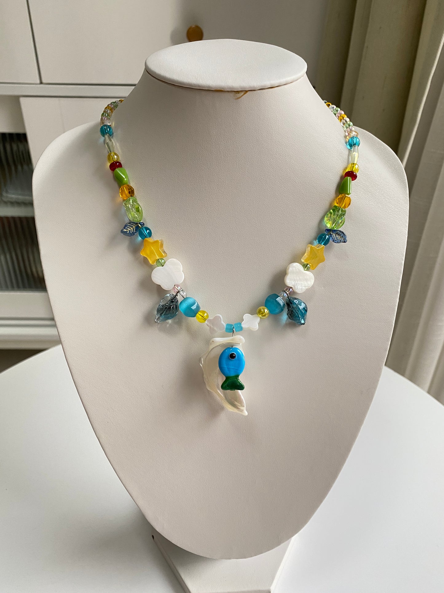 One-of-one handmade shell necklace displayed on a necklace bust, featuring a blue-green Czech glass fish pendant and natural coastal-inspired details.
