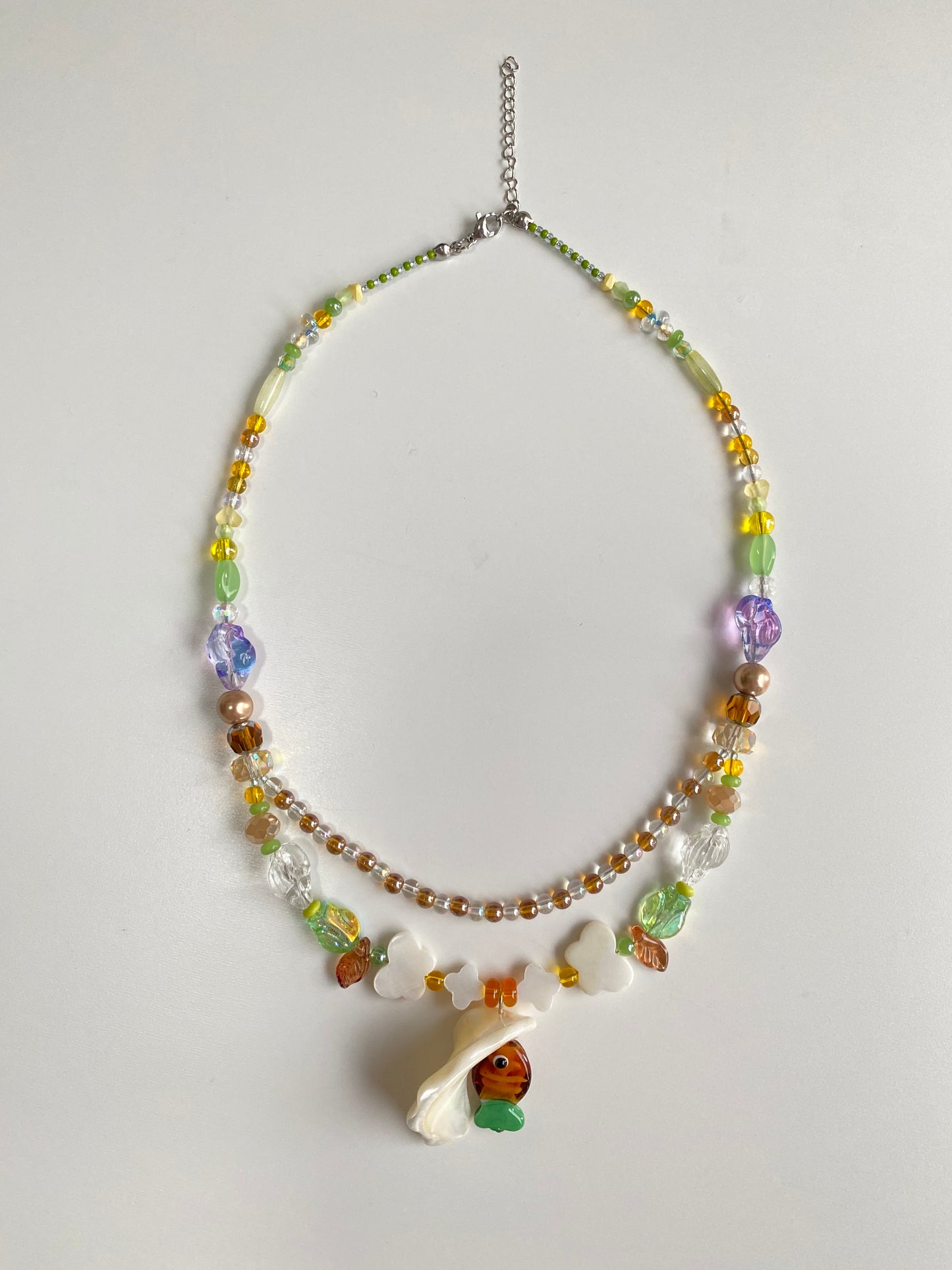 Close-up of a handmade layered necklace featuring a brown-green glass fish, translucent bubble bead strands, butterfly shell charms and glass conch accents.