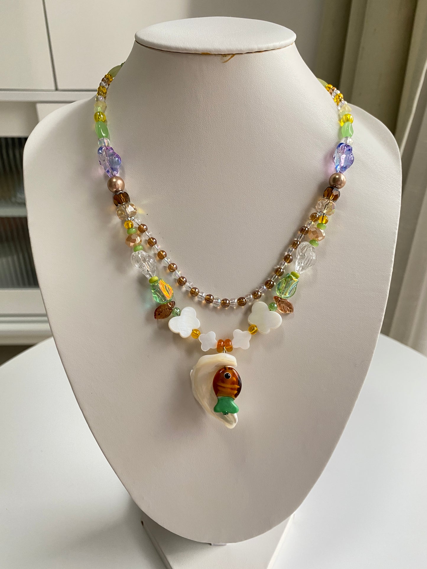 Handcrafted ocean necklace displayed on a necklace bust with a natural shell pendant, colorful Czech glass fish and layered coastal-inspired beadwork.