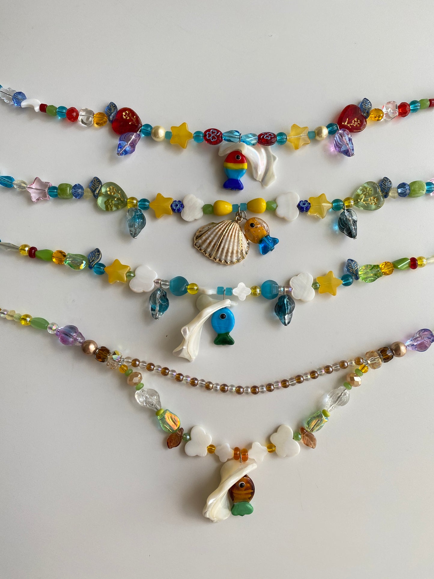 Close-up of four handmade beach necklaces showcasing colorful glass fish pendants, seashell details, butterfly shell accents and sparkling seed beads.